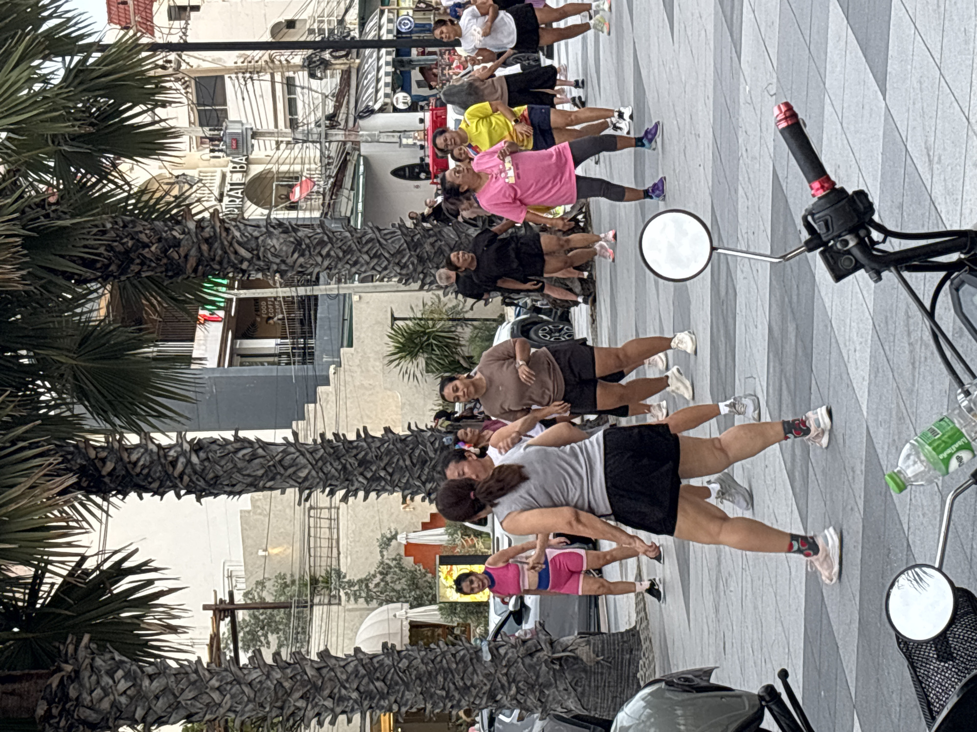 A group of approximately 15 to 20 individuals, predominantly women, are engaged in an outdoor exercise class on a tiled walkway in Pattaya City, Thailand. The scene is framed by tall palm trees on either side, with a street and buildings visible in the background. The participants are dressed in athletic wear, including shorts and t-shirts of various colors such as pink, yellow, white, and brown. Some are actively moving, with raised arms and bent knees, suggesting coordinated movements. A motorcycle is partially visible in the foreground, with its handlebars, mirror, and a plastic water bottle on the ground. The environment appears to be a public area, possibly a promenade or sidewalk adjacent to shops and other establishments, as indicated by the architectural style of the buildings and the presence of streetlights. The lighting suggests it is daytime, possibly late afternoon or early evening.