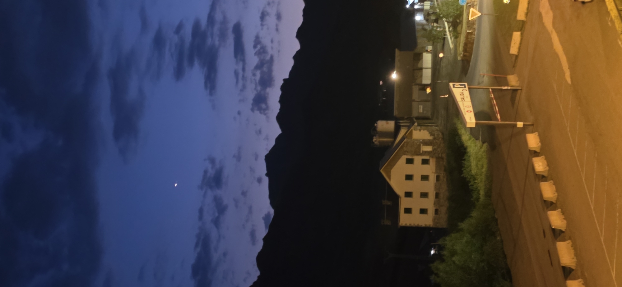 The image depicts a twilight scene in Sallent de Gállego, Spain. In the foreground, a paved area with white parking space markings and concrete barriers is visible. A sign with the word "SALIDA" (Exit in Spanish) and a parking symbol is centrally located. Behind this, a two-story building with a light-colored facade, stone accents, and numerous square windows is present, alongside other industrial-looking structures with visible tanks. In the background, dark, silhouetted mountains rise against a gradient sky transitioning from deep blue to lighter shades, with scattered clouds. A sliver of a crescent moon is visible in the upper left portion of the sky. The overall atmosphere is serene and somewhat industrial.
