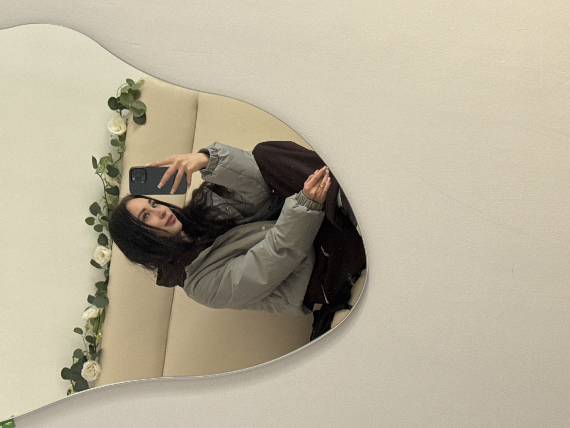 The image captures a mirror selfie of a woman taken indoors. The mirror itself has an irregular, wavy-edged shape, reflecting the woman and a segment of the room. The woman has long, dark hair and is wearing a light grey or sage-colored puffer jacket paired with dark-colored pants. She holds a dark smartphone in her left hand, appearing to be taking the picture, and a subtle ring is visible on her right hand.
Behind her in the reflection is a light beige or cream-colored padded surface, possibly part of a booth seat or a decorative wall panel. A vertical floral garland featuring green leaves and several white roses runs along the left side of the reflection, adding a decorative touch to the environment. The visible parts of the room outside the mirror's reflection consist of a plain, light-colored wall.
The woman is looking towards the camera, likely seated or leaning back in a relaxed posture. The overall atmosphere suggests a contemporary indoor setting, possibly a cafe, restaurant, or lounge, characterized by a clean and light aesthetic. The lighting appears soft and uniform, typical of an indoor space. There is no visible text in the image. The image is displayed rotated 90 degrees counter-clockwise from a natural upright perspective.