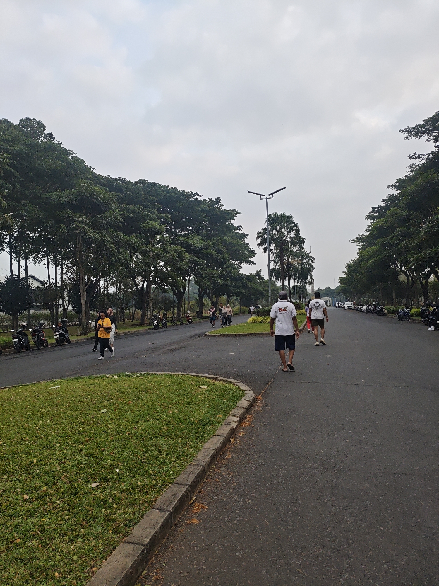 The image depicts a cloudy day on a paved road lined with trees and a grassy median in Bacin, Indonesia. Several individuals are walking or jogging on the road. On the left side of the road, a row of parked motorcycles is visible. In the foreground, a curve of grass and a concrete curb separate the road from a green lawn. A street lamp is situated further down the road.