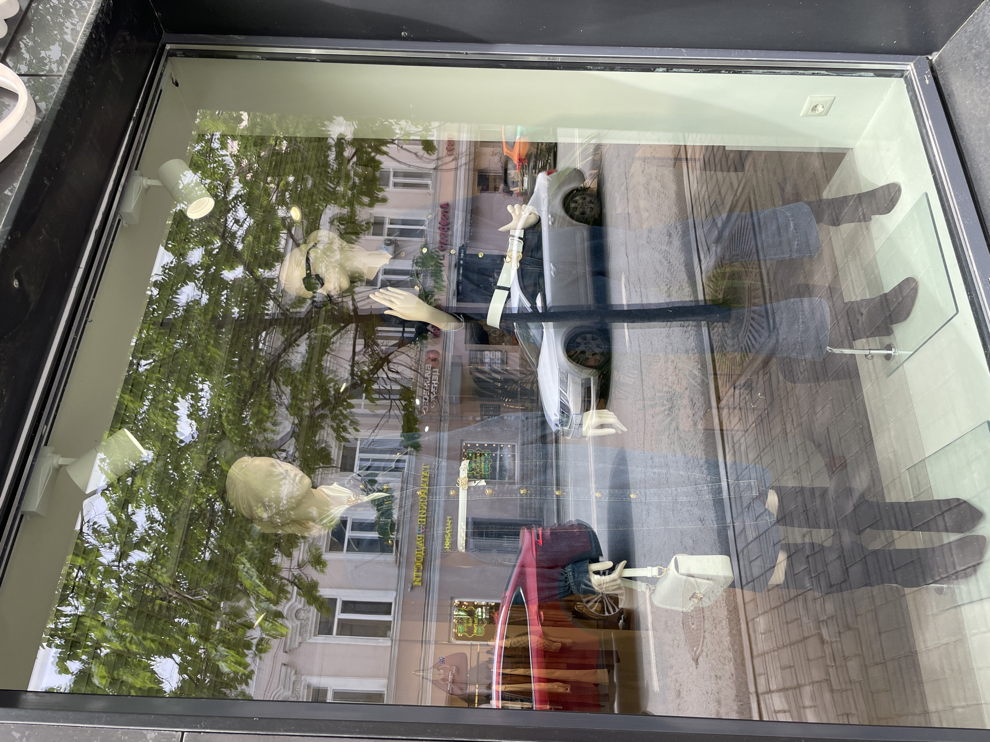 The image shows a storefront window display in Kazan, Russia. Reflected in the window is a building facade with windows and signage, along with trees and a white car. Inside the window, there are two headless mannequins wearing clothing. The mannequins are arranged vertically, with one positioned near the top of the window and the other near the bottom. The window interior has a light fixture on the upper left and an electrical outlet on the upper right. A purse sits next to the bottom mannequin. The overall scene is a blend of the interior display and the reflections of the exterior environment.