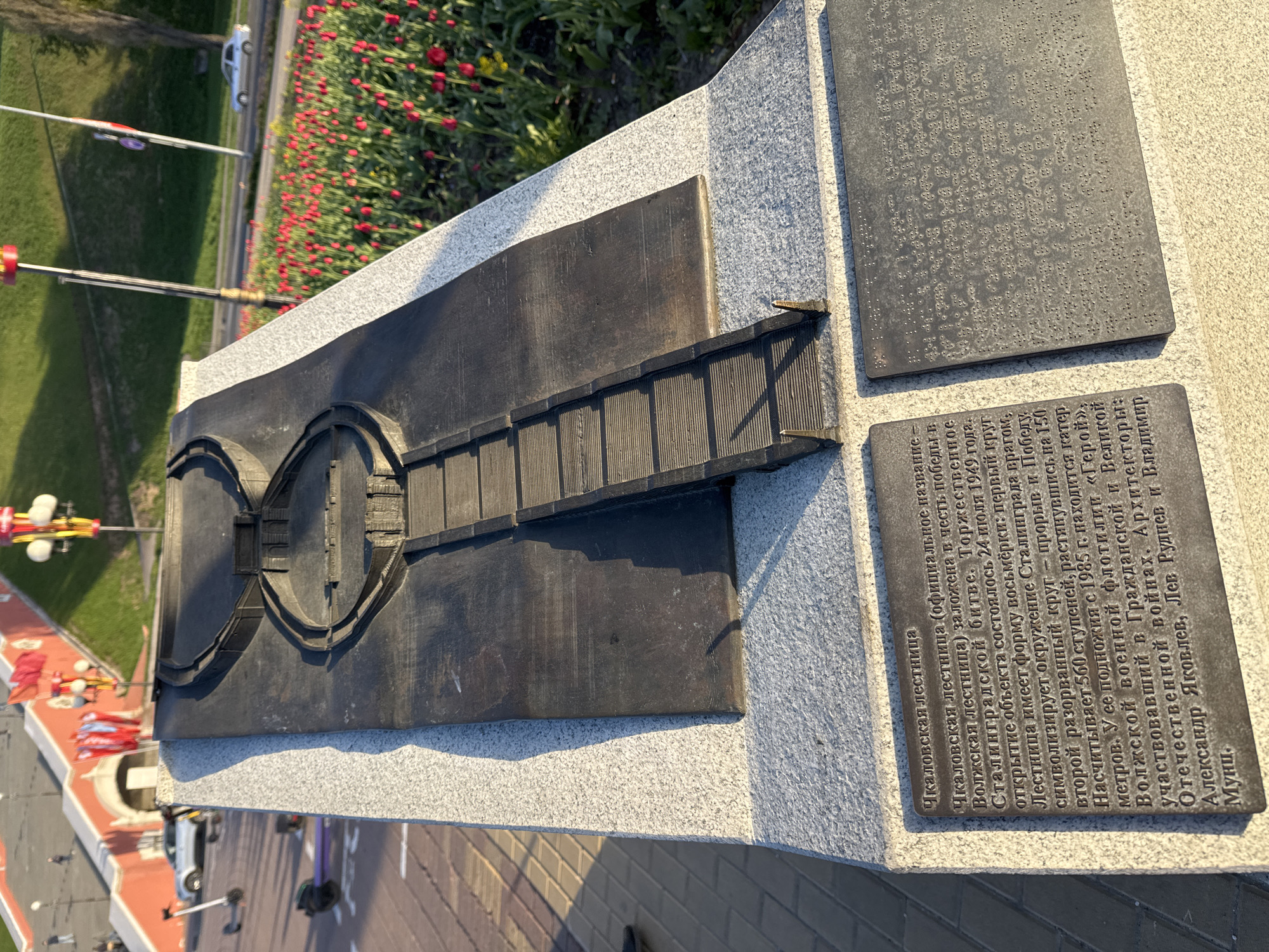 The image captures a detailed view of a memorial dedicated to the Chkalov Staircase, also known as the Volzhskaya Staircase, in Nizhny Novgorod, Russia. The memorial is made of stone. It features a bronze sculpture of the staircase itself, depicting its distinctive figure-eight shape, symbolizing the encirclement of Stalingrad by enemy forces, and the subsequent breakthrough representing victory. Adjacent to the sculpture are plaques with text in Russian, providing historical context and information about the staircase's significance, its construction, and its dedication to the victory in the Battle of Stalingrad. The image also captures a glimpse of the surrounding environment, including greenery, other parts of the memorial, and hints of the urban landscape.