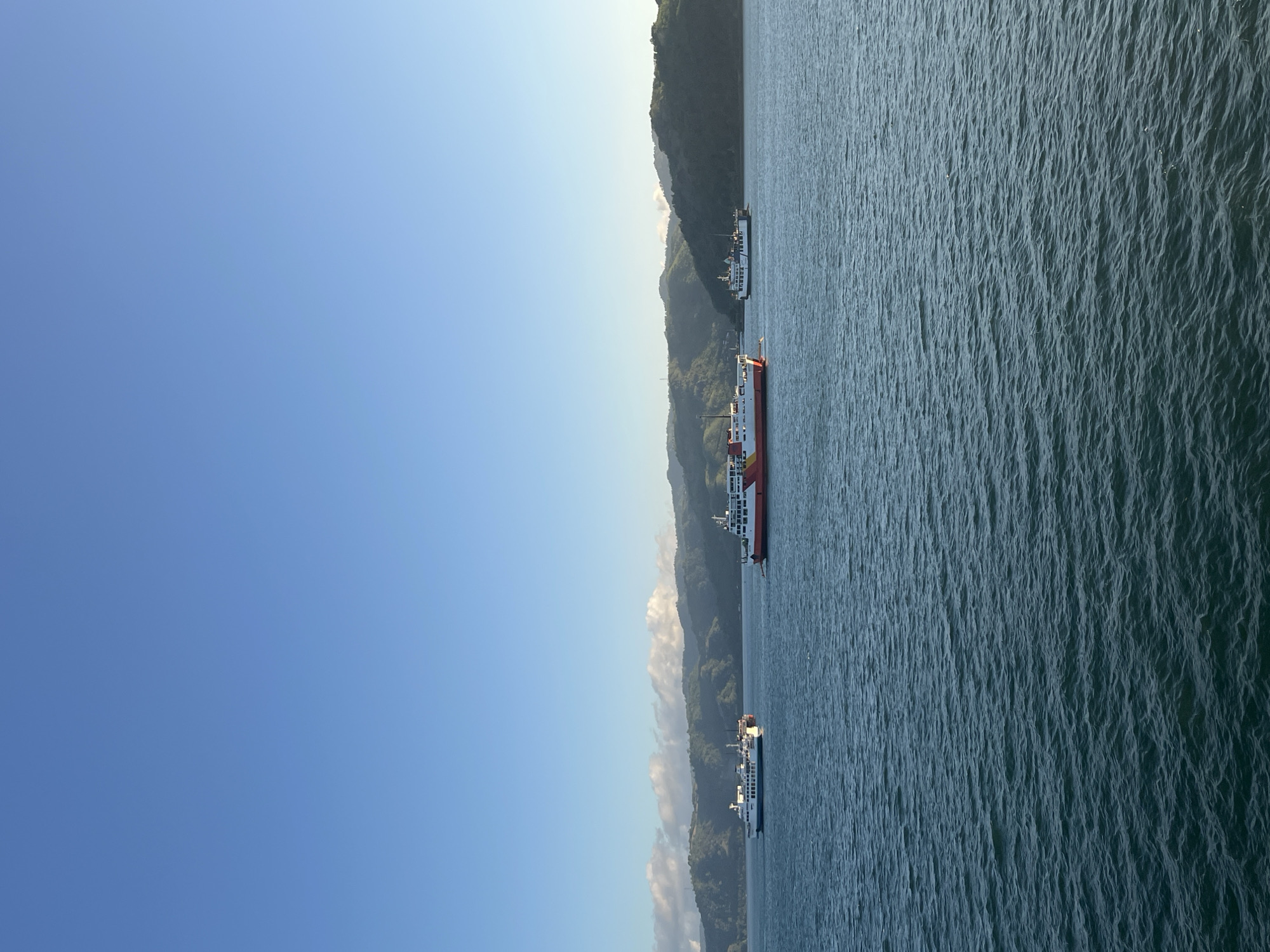 The image depicts a serene seascape with three ferries navigating the waters. The ferries are positioned at varying distances from the viewer, with the closest one being a red and white vessel, followed by a white ferry with blue accents, and a third white ferry further in the distance. The water is calm with gentle ripples. In the background, lush green hills and mountains rise, partially covered by wispy clouds. The sky is clear and a vibrant blue, suggesting a pleasant day. The location is identified as Batas Desa Skotong Timur, Indonesia. There are no people visible in the image.