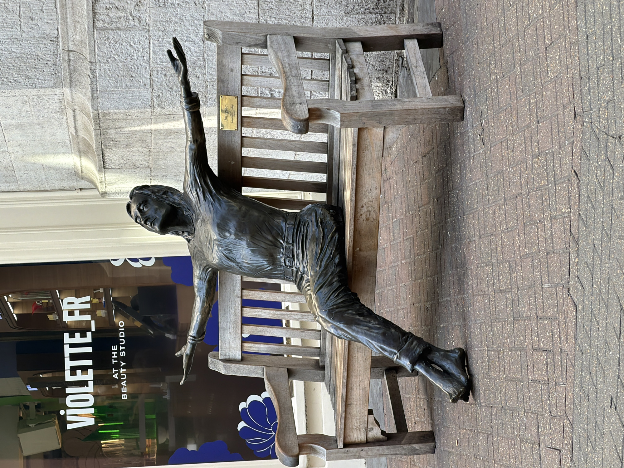 The image depicts a bronze sculpture of a seated woman with her arms raised, placed on a wooden bench in front of a shop called "VIOLETTE FR at the Beauty Studio". The sculpture is positioned so that the woman appears to be interacting with the environment. The bench is situated on a brick paved surface. The location is in the city of London, United Kingdom, as the shop name "VIOLETTE FR" is the name of a beauty studio that has a location in London.