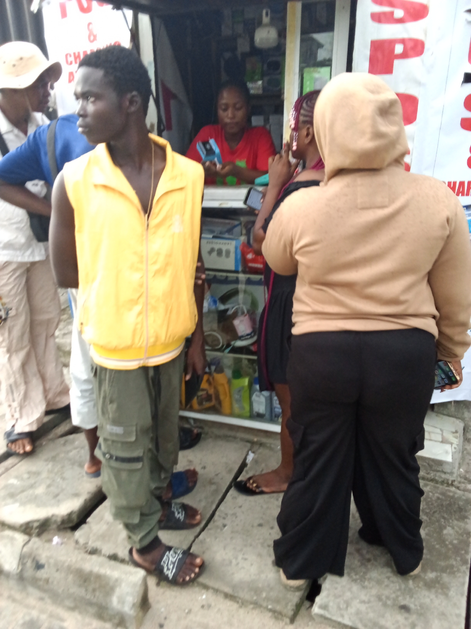 This image captures a street scene in Ado, Nigeria, depicting several individuals gathered outside a small, open-front shop.
In the foreground, slightly left of center, a dark-complexioned male stands facing right, looking slightly downwards. He wears a bright yellow sleeveless zippered vest over a dark garment or bare chest, olive green cargo pants, and black-and-white slide sandals. A gold-colored chain is visible around his neck, and he holds a dark object in his right hand by his side.
To his right, facing away from the viewer, a person with a dark complexion wears a light beige hooded sweatshirt with a textured pattern and loose-fitting black pants, ending in light-colored closed-toe shoes. This individual holds a smartphone with a gold-colored case in their left hand, held low by their side.
Between these two, partially obscured, is another dark-complexioned individual, possibly a female, wearing a dark top and sporting reddish-purple braided hair. This person faces left, with their left hand raised near their face.
Inside the shop, positioned behind a counter, a dark-complexioned female wearing a red short-sleeved shirt is seated. She is looking down, holding a blue object in her hands. The shop's interior features shelves stocked with various items, including electronics and bottled liquids. Partially visible signs on the shop's exterior include red letters "AP" and text indicating "FU & CHARGING."
To the far left of the frame, another dark-complexioned male stands, partially obscured. He wears a light-colored button-up shirt, a light-colored wide-brimmed hat, and light-colored loose pants with dark sandals. The legs and feet of an additional individual wearing white shorts and a light brown sandal are visible between this man and the man in the yellow vest.
The ground is concrete, showing minor cracks and unevenness. The scene appears to be set during daylight hours.