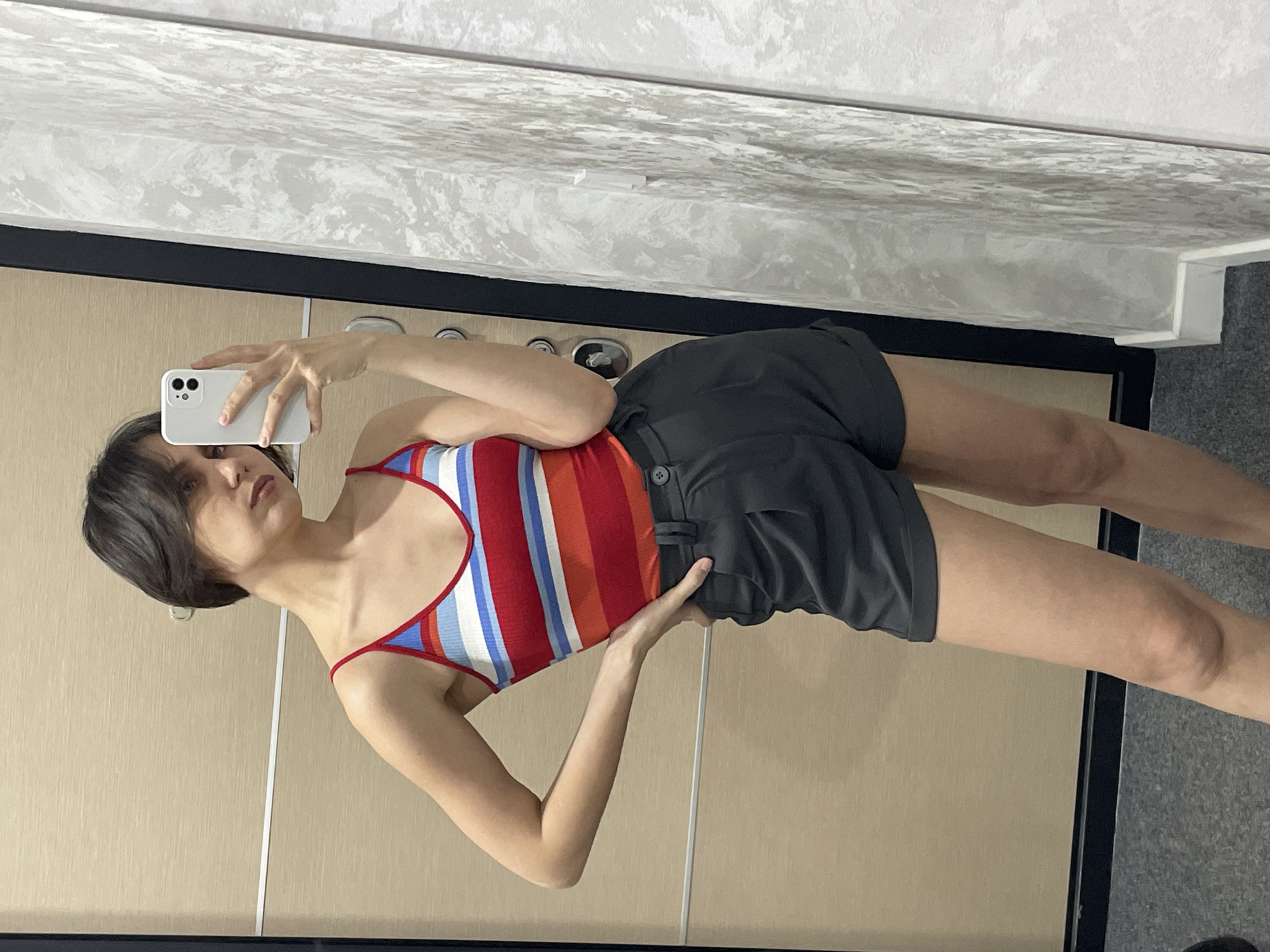 A woman stands indoors, facing a mirror and holding a white smartphone to take a selfie. She is wearing a striped red, white, and blue top with thin red straps and dark gray shorts. The background includes a mirrored door with vertical panels and a ceiling with a textured, possibly marbled, pattern. The floor is gray. It is impossible to establish the location in the city of Izhevsk, Russia from the provided image.