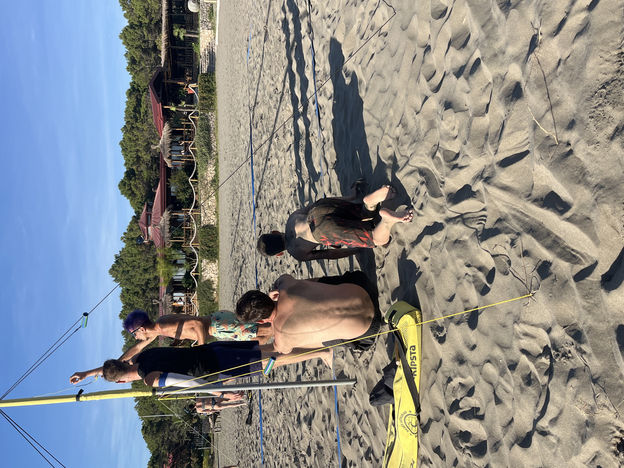 The image depicts a sandy beach scene, likely in Pistula, Montenegro, given the context of the city. The foreground is dominated by sand dunes and shadows cast by overhead structures. In the mid-ground, four individuals are present on the beach. Two men are standing near a yellow volleyball pole, one of whom appears to be adjusting a string or rope attached to the pole. Another man is sitting on the sand, facing away from the camera, with visible tan lines on his back. A younger individual is kneeling on the sand in front of the sitting man, also facing away. A yellow volleyball bag with the brand name "KIPSTA" is visible on the sand in the lower left quadrant of the image. In the background, a row of trees and several thatched-roof buildings suggestive of a beachfront establishment are situated behind a stone wall. A woman in a bikini is partially visible walking in the distance. The sky is clear and blue.