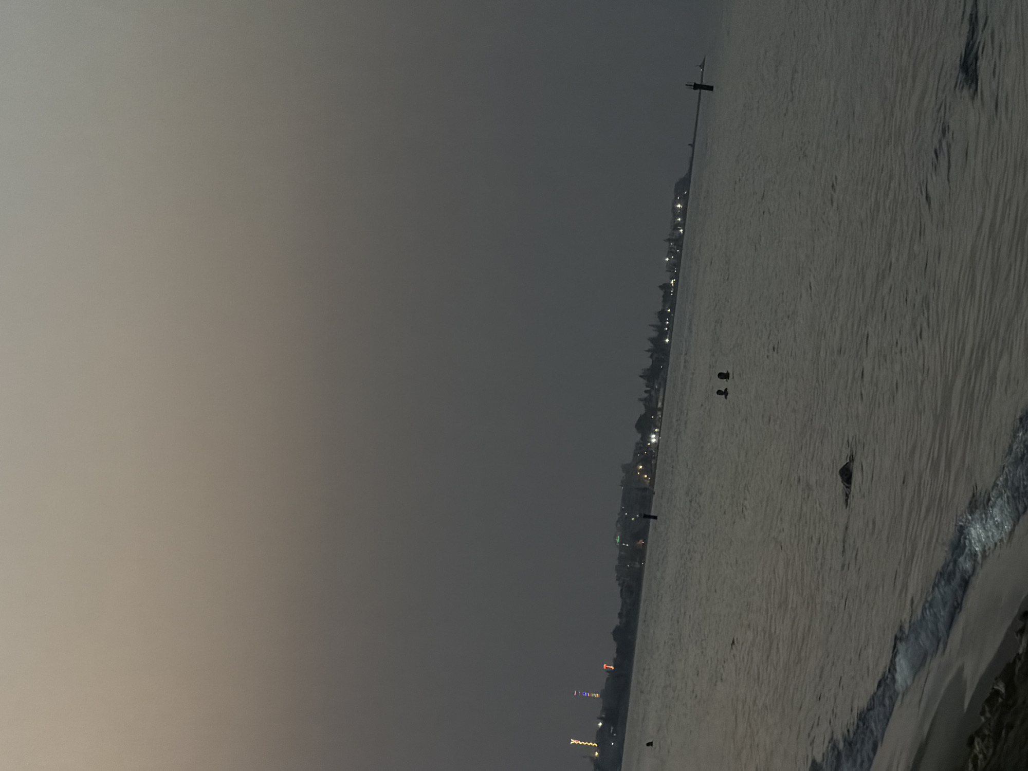 The image depicts a twilight scene at a beach in Sousse, Tunisia. The sky is overcast and a muted gray, suggesting either dusk or dawn. In the distance, a shoreline is visible with a scattering of buildings and lights, indicating an urban or developed coastal area. Several illuminated structures, possibly decorative lights or towers, stand out along the coastline. The sea is calm with gentle ripples and waves lapping at the sandy shore in the foreground. Several figures are present in the water; two individuals appear to be swimming or wading near the middle ground, and another dark shape, possibly an animal or person partially submerged, is visible closer to the shore. A black buoy or marker stands upright in the water further out. The overall atmosphere is serene and quiet, typical of a beach at the edge of the day.