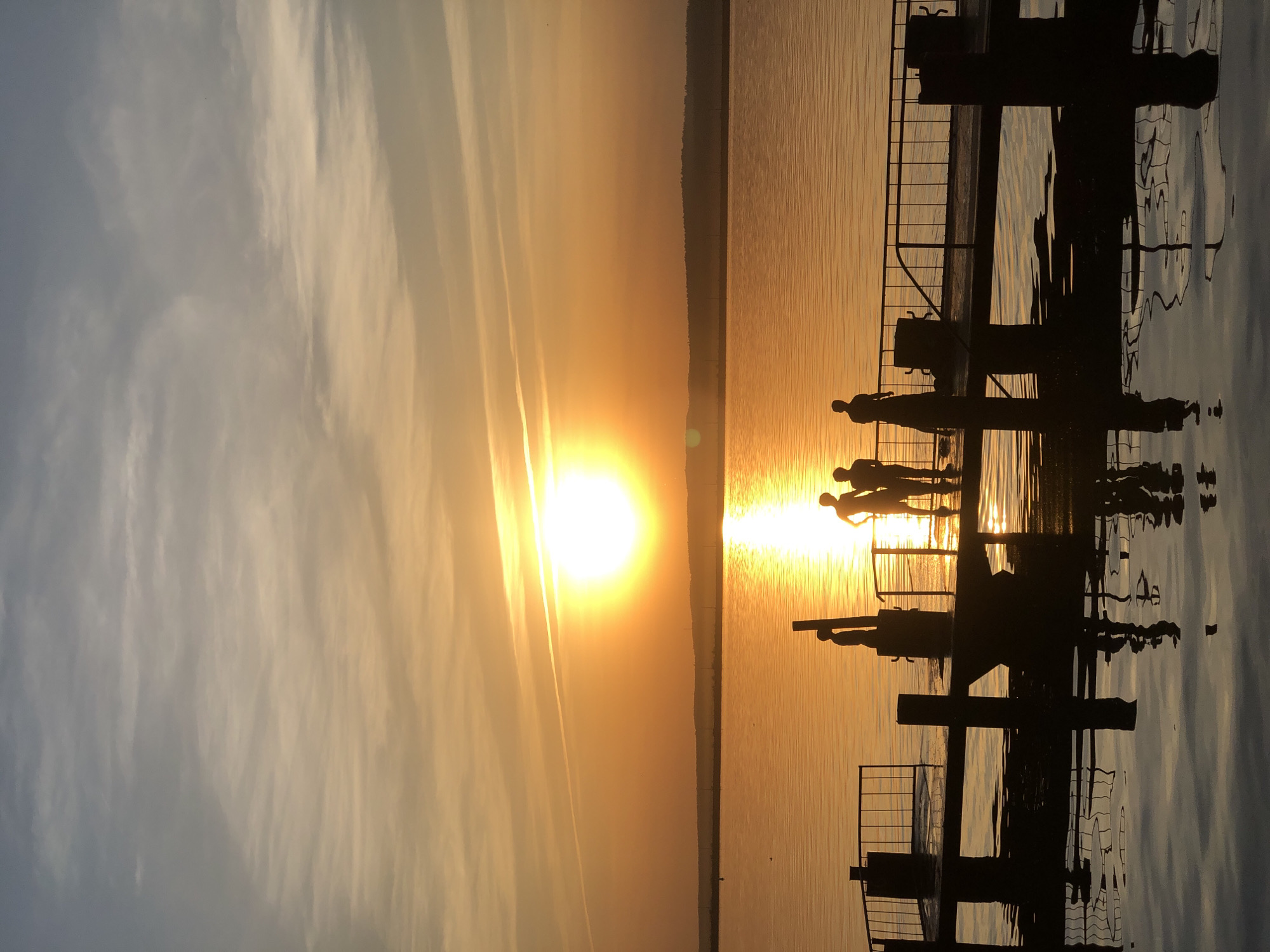 The image shows a sunset scene, likely in Chaikovsky, Russia, over water. The sun is positioned near the horizon, casting a bright glow and creating a silhouette effect on a pier or dock structure in the foreground. Several people are standing on the pier, their figures appearing as dark silhouettes against the bright water. The pier has railings and other structural elements. The water reflects the sunlight, and the sky transitions from lighter hues near the sun to darker colors further away. Some clouds are visible in the sky.