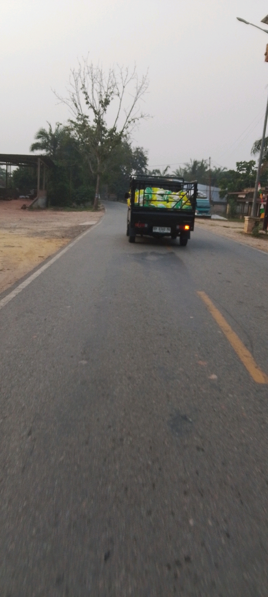 A black pickup truck with a metal cage bed is driving on a paved road. The truck bed is filled with several large, light-colored sacks, some of which are yellow. The truck's license plate is visible and reads "BP 8388 PN". The road has a broken yellow line painted down the center. On the left side of the road, there is a dirt area with some structures, possibly a building with an open roof. There are trees and vegetation on both sides of the road. The sky is overcast and light. The scene appears to be in a rural or semi-rural area.