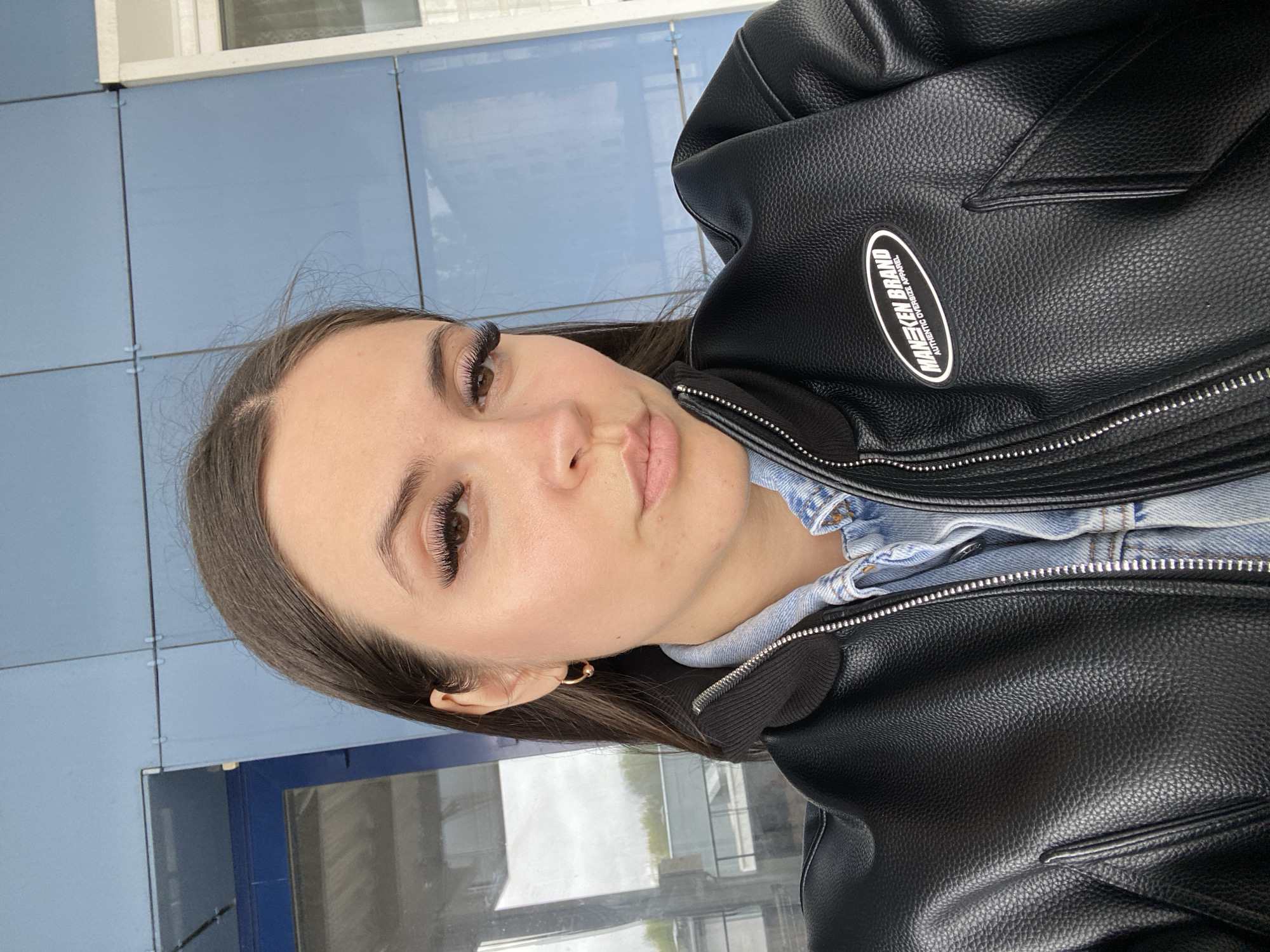 A woman is posing for a photograph in front of a building with blue tile panels. She is wearing a black leather jacket over a light blue denim shirt. The jacket has a logo patch that reads "MEECKEN BRAND AUTHENTIC OVERSIZE APPAREL." She is facing the camera and is wearing false eyelashes and small gold hoop earrings. The location appears to be in an urban area in Chaikovsky, Russia, as suggested by the building architecture and style.