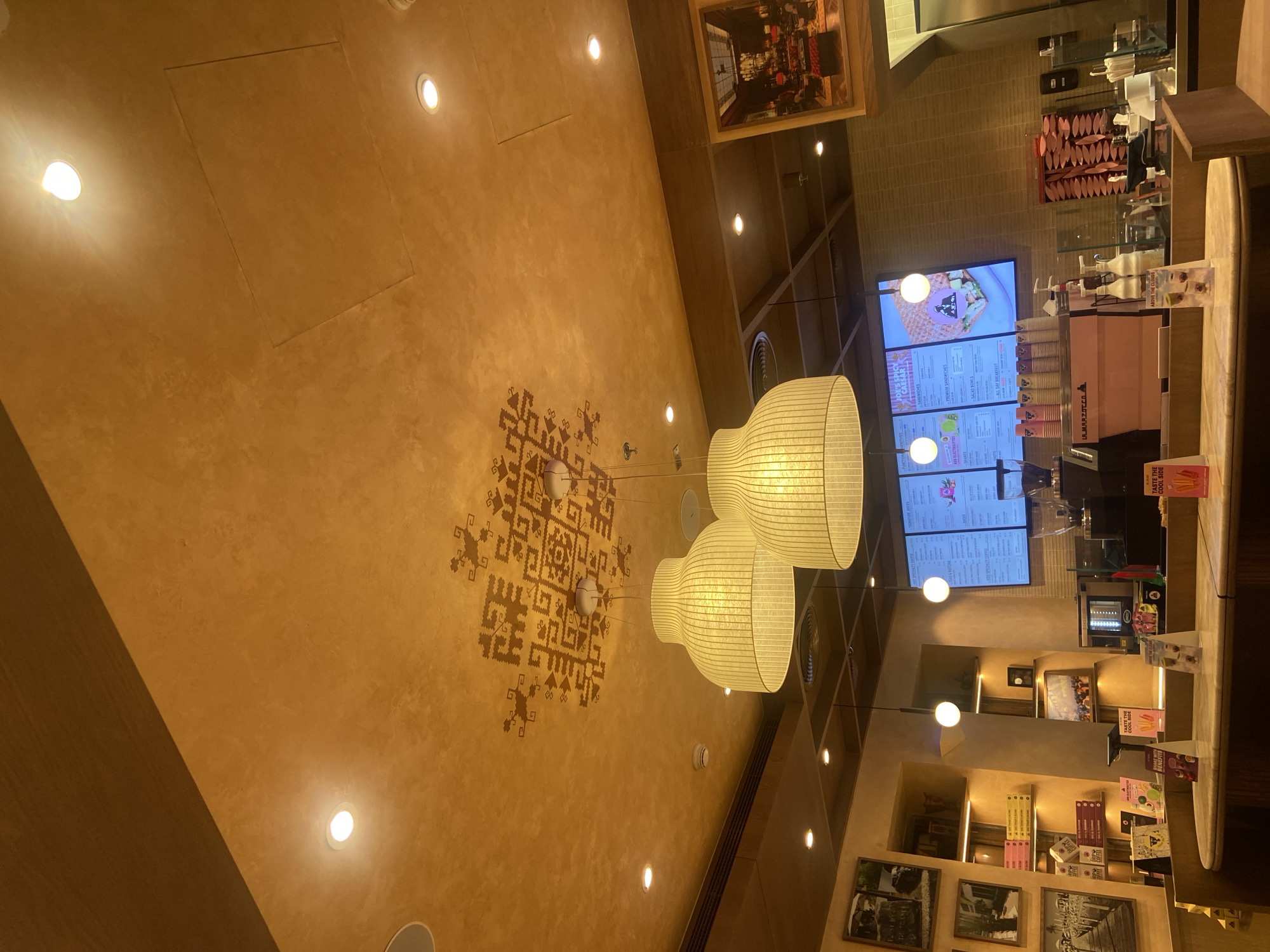 This image depicts the interior of a cafe in Deira, United Arab Emirates. The ceiling is a textured ochre color with a geometric pattern in a darker shade. Two large, bell-shaped pendant lamps with ribbed shades hang from the ceiling, emitting a warm yellow light. Several recessed spotlights are also visible on the ceiling.
The cafe counter is prominent in the foreground, made of a light-colored curved material. Behind the counter, a digital menu board displays various food and drink options, including sandwiches and smoothies. A coffee machine, likely a La Marzocco, is positioned on the counter, along with dispensers for syrup and cream. Several stacks of colorful cups are also visible.
To the left of the counter, shelves are lined with boxed products, some appearing to be coffee or food items. Framed pictures hang on the wall above the shelves, depicting scenes that seem to be of outdoor locations. A framed artwork hangs on the wall to the right, showing an interior scene with people and furniture.
The overall ambiance is warm and inviting, with a focus on a modern cafe aesthetic. The lighting creates a cozy atmosphere, and the decor suggests a contemporary establishment. No people are clearly visible in the frame.