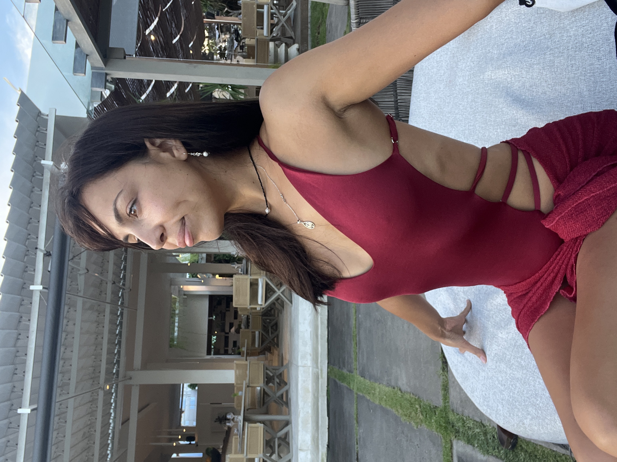 An outdoor, eye-level shot captures a woman seated at what appears to be an outdoor restaurant or resort area in Kuta Selatan, Indonesia. The woman, with dark hair and a gentle smile, is looking towards her left. She wears a dark red one-piece swimsuit or bodysuit, featuring strap details on the side, paired with a matching textured red fabric that resembles a skirt or wrap. Her accessories include pearl drop earrings and two necklaces: a black beaded choker and a silver chain with a small, indistinct pendant.
Her right arm is visible, with her hand resting on a light-colored table surface. The background features modern, light-toned architecture, including slatted roofs or pergolas, and outdoor dining furniture like wooden chairs and tables. Patches of green grass are visible on the ground. The lighting suggests late afternoon or early evening, casting soft light and indicating clear, pleasant weather. No visible text is present in the image. The overall atmosphere conveyed is relaxed and serene.