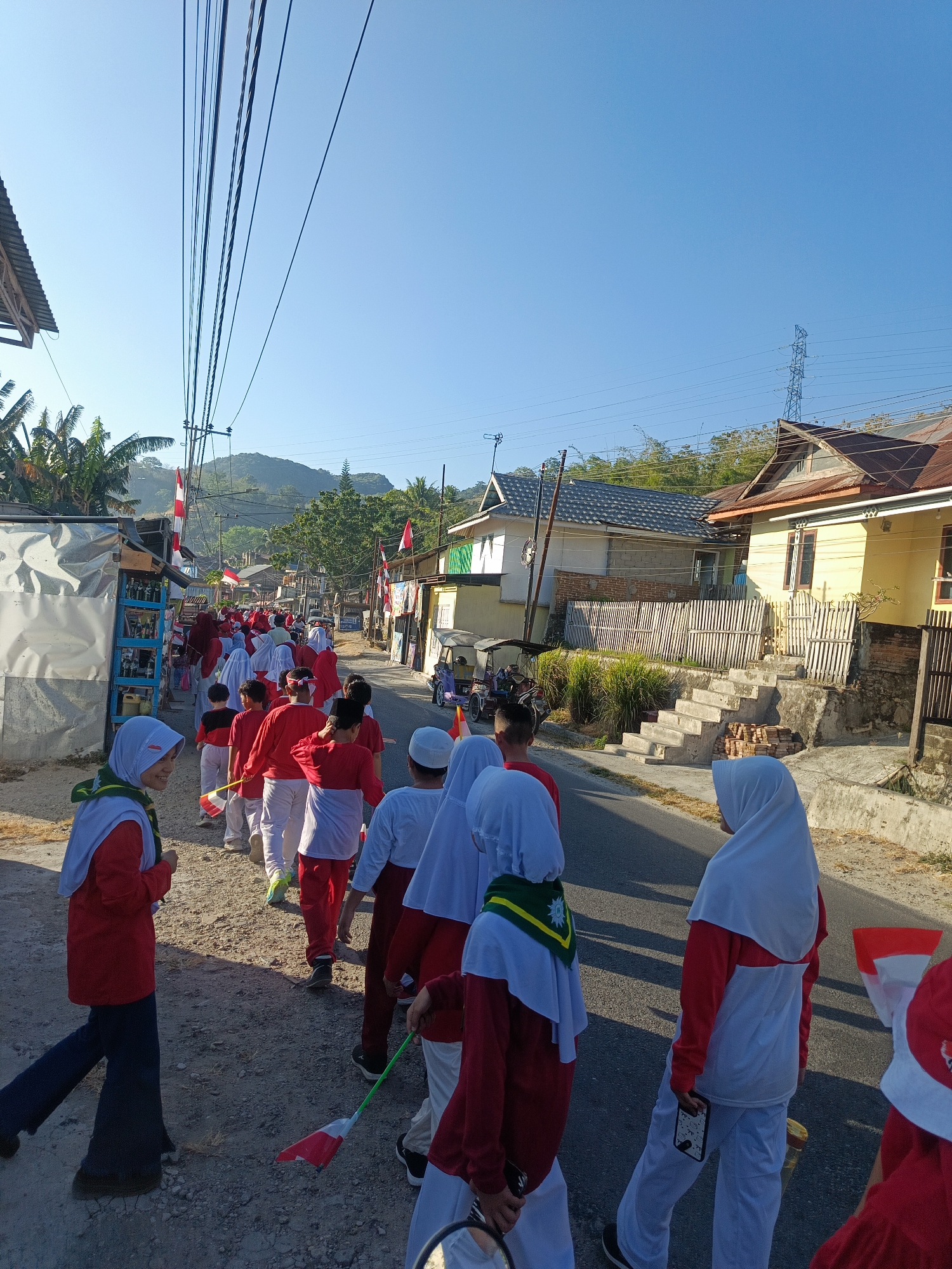 A procession of children, mostly wearing red and white attire, walks down a dusty street lined with houses and utility poles in Gorontalo, Indonesia. The children are carrying small Indonesian flags, suggesting a national celebration. The sky is clear blue and the sun is bright, casting shadows on the ground. Some of the children are wearing hijabs. The houses appear to be modest, with some featuring tin roofs. The street is paved with asphalt in the distance but is dusty and uneven where the children are walking. In the background, a power line tower is visible on a hillside. The scene depicts a local community participating in a patriotic event.