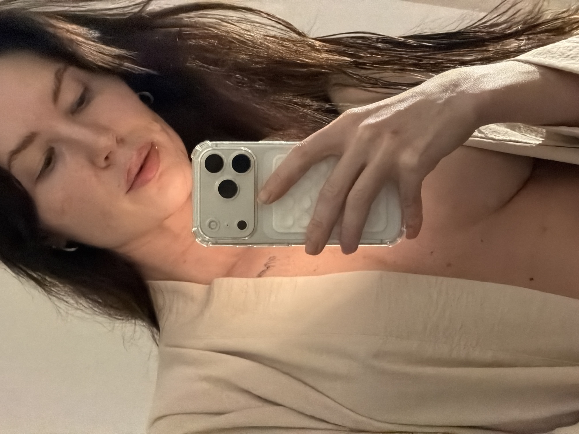 A selfie-style image shows a woman holding a smartphone, obscuring part of her upper body. She has long, dark hair and fair skin. The woman's face is visible in the left side of the frame. She is wearing a light-colored robe or shirt. The location is not discernible from the image.