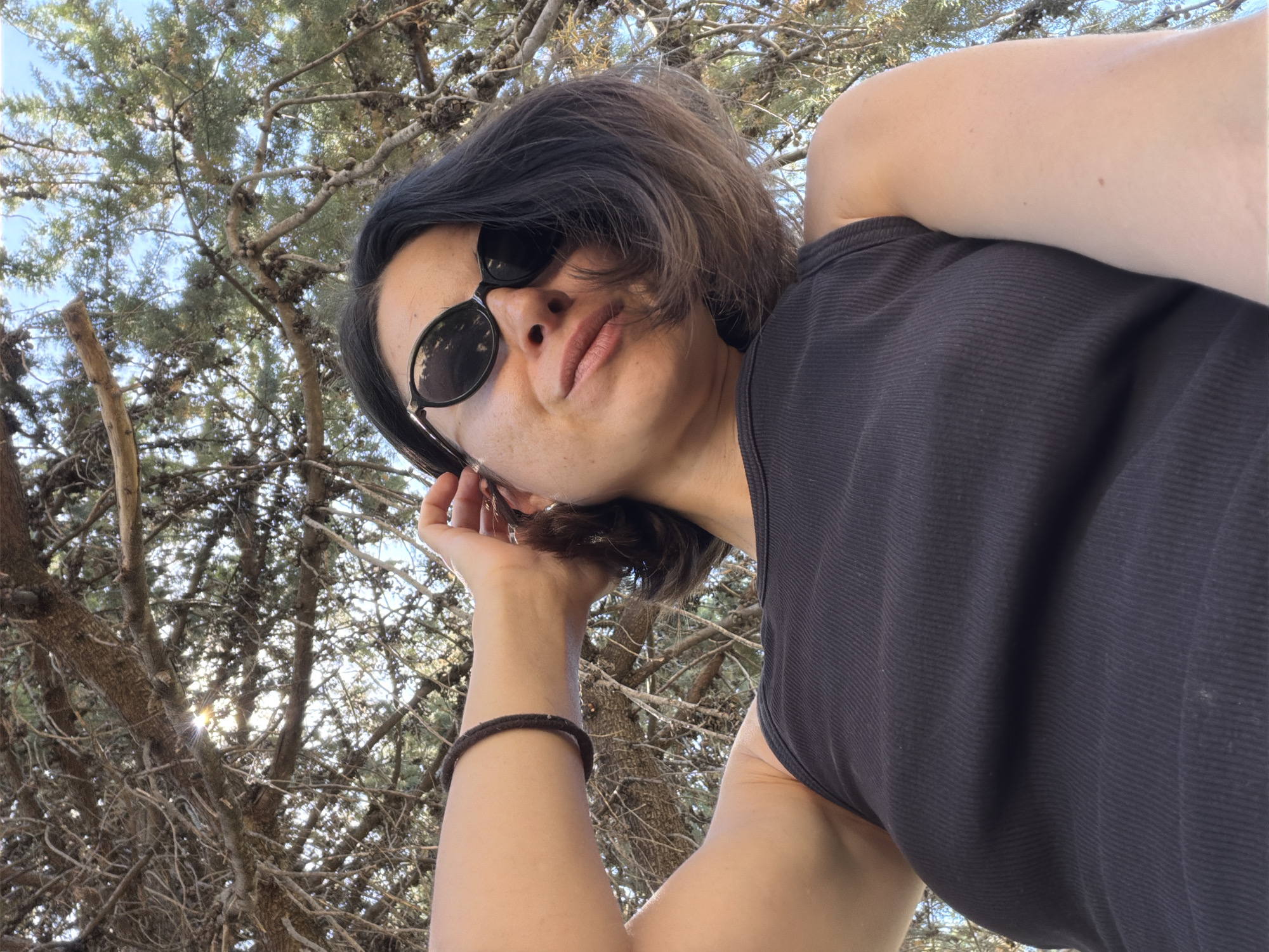 The photograph depicts a woman wearing a black tank top and sunglasses, posing with her head tilted back. She is wearing a bracelet on her left wrist and is touching her hair with her left hand. The background shows tree branches and a blue sky with some sunlight. The image was taken outdoors, possibly in a park or wooded area. There is no information to definitively identify the location as Budva, Montenegro, beyond the request for the location.