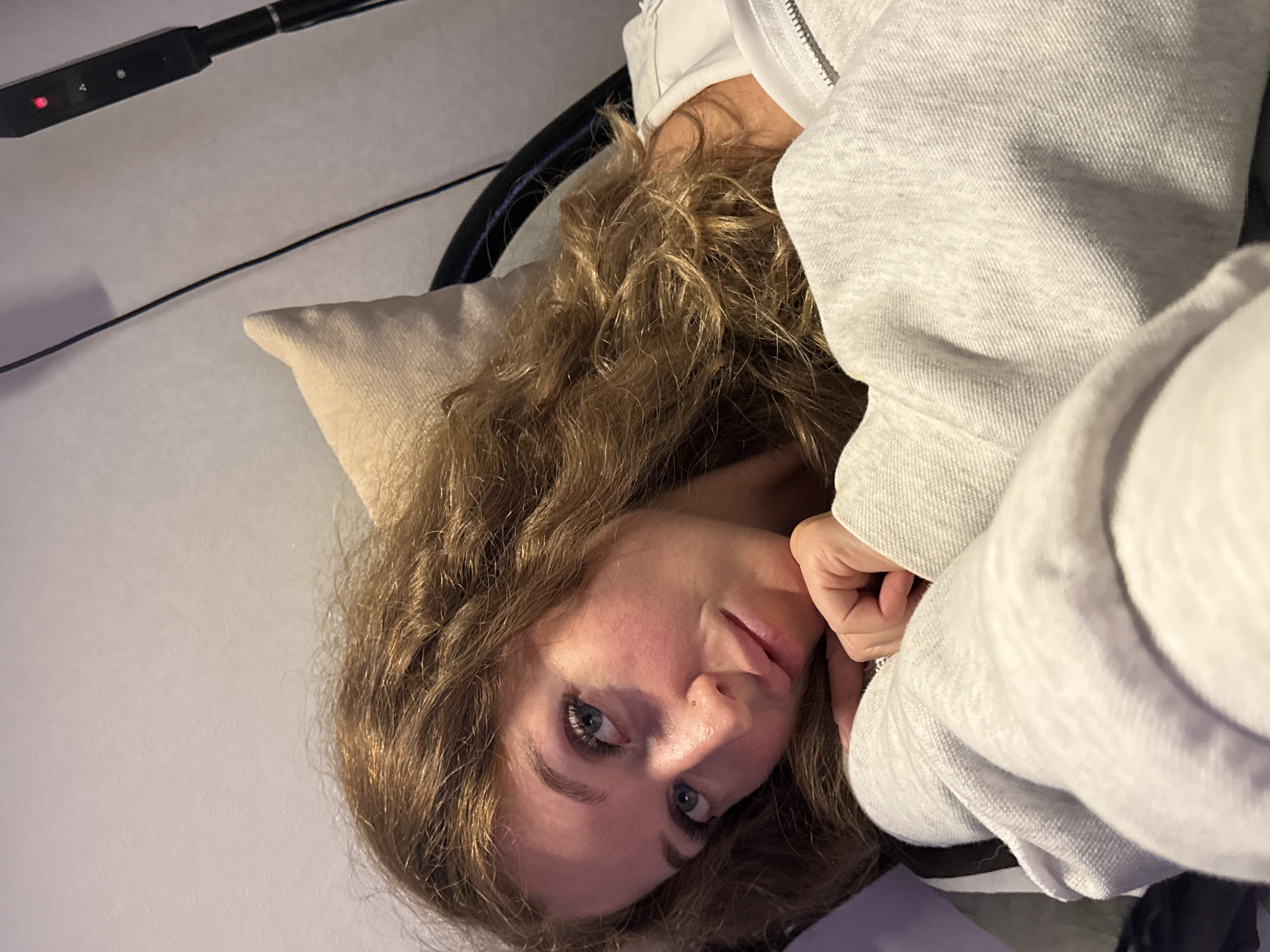 A fair-skinned woman with curly, light brown hair and blue eyes is looking directly at the camera. She is wearing a light gray hooded sweatshirt. She is lying down on a light tan pillow. A black object with buttons is visible in the upper left corner, along with a black cord. The background is a light-colored wall. There is no information to indicate that the image was taken in the city of Chaikovsky, Russia.