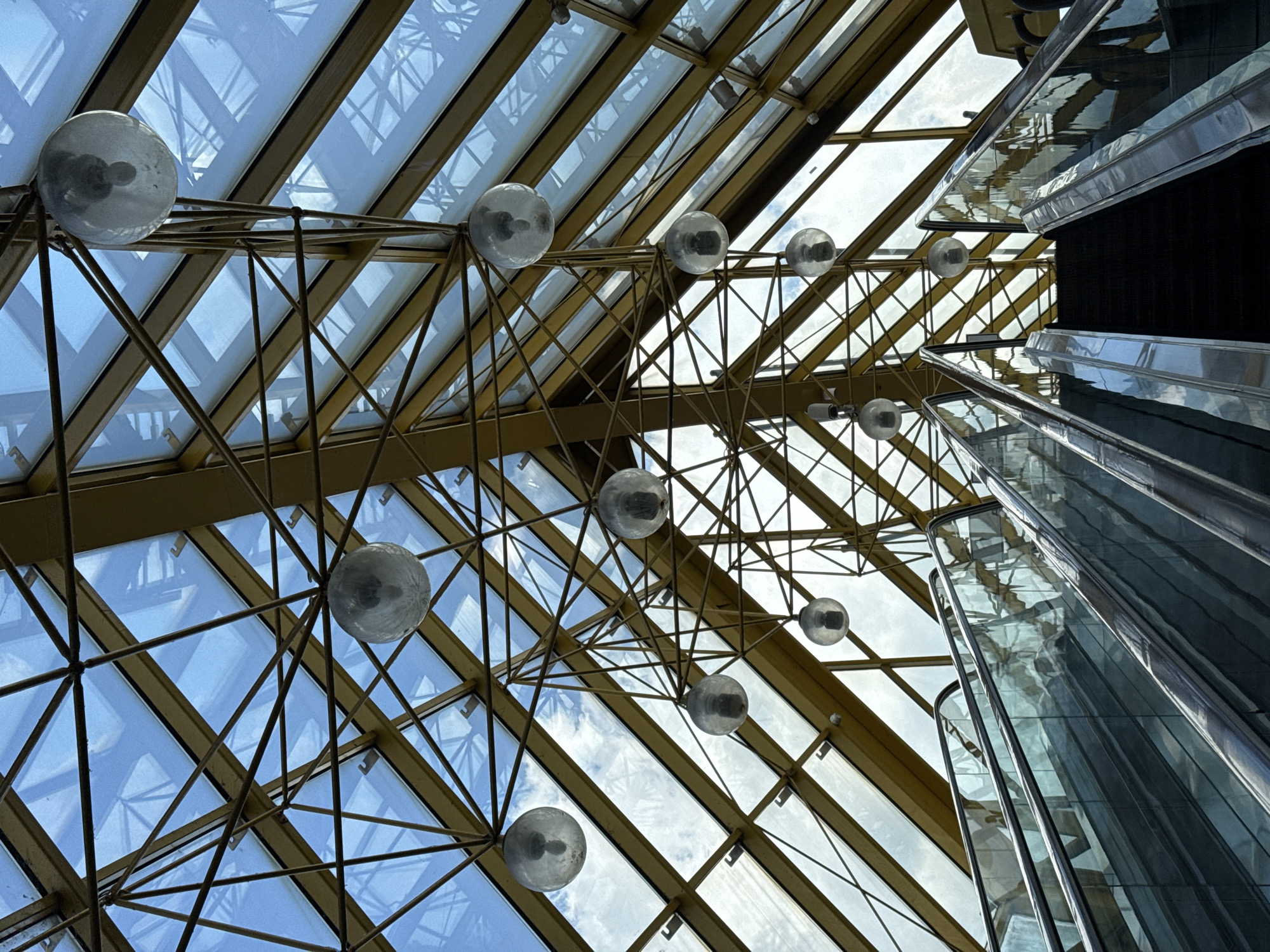 An upward-angled shot captures the architectural details of a large indoor space, likely an atrium or mall. The dominant feature is a massive glass ceiling or roof, structured with a complex lattice of golden-colored metal beams and frames, forming numerous triangular and rectangular panes. Through these clear glass panels, a bright, partly cloudy daytime sky is visible.
Suspended from the golden framework are multiple spherical light fixtures, each containing a visible light bulb filament. These lights are scattered across the ceiling, adding to the geometric complexity of the scene.
On the right side of the frame, a modern glass escalator or moving walkway ascends vertically. Its polished metal and glass surfaces reflect the ceiling structure and the ambient light, creating abstract patterns. The design suggests a contemporary public building. There are no people visible, and no specific activity is taking place other than the structural presence of the architecture itself. No visible text is present in the image.