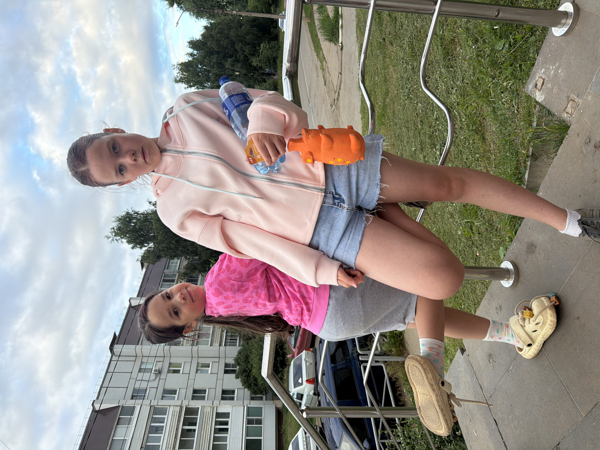 Two young girls are posing for a photo outdoors, on a cloudy day. The girl in the foreground, on the right, is wearing a pink hoodie, denim shorts, white socks with colorful polka dots, and beige clogs. She is holding an orange toy and a bottle of water. She has dark hair and is smiling. The girl behind her, on the left, is wearing a pink sweatshirt with a heart pattern and grey shorts. She has brown hair pulled back and is looking at the camera with a neutral expression. The background shows a modern apartment building, trees, and a metal railing. The scene appears to be a residential area in Chaikovsky, Russia.