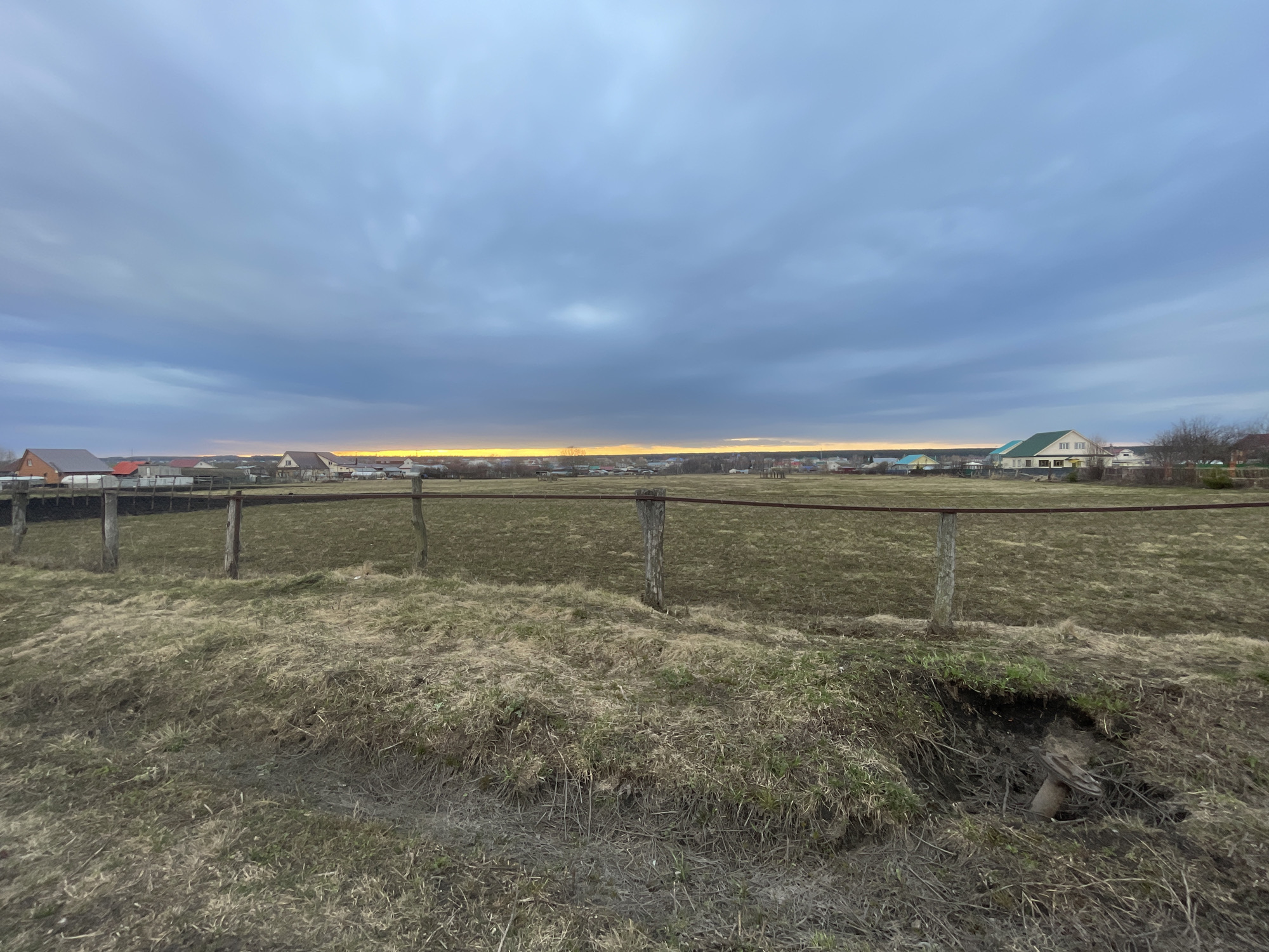 The image shows a rural landscape in Новая Шемурша, Russia, under a cloudy sky with a hint of sunset on the horizon. In the foreground is a ditch with dry grass and bare earth. Beyond the ditch, there's a field enclosed by a rustic fence made of wooden posts and a single horizontal rail. In the distance, a line of houses is visible. There are no people or animals evident in the image.