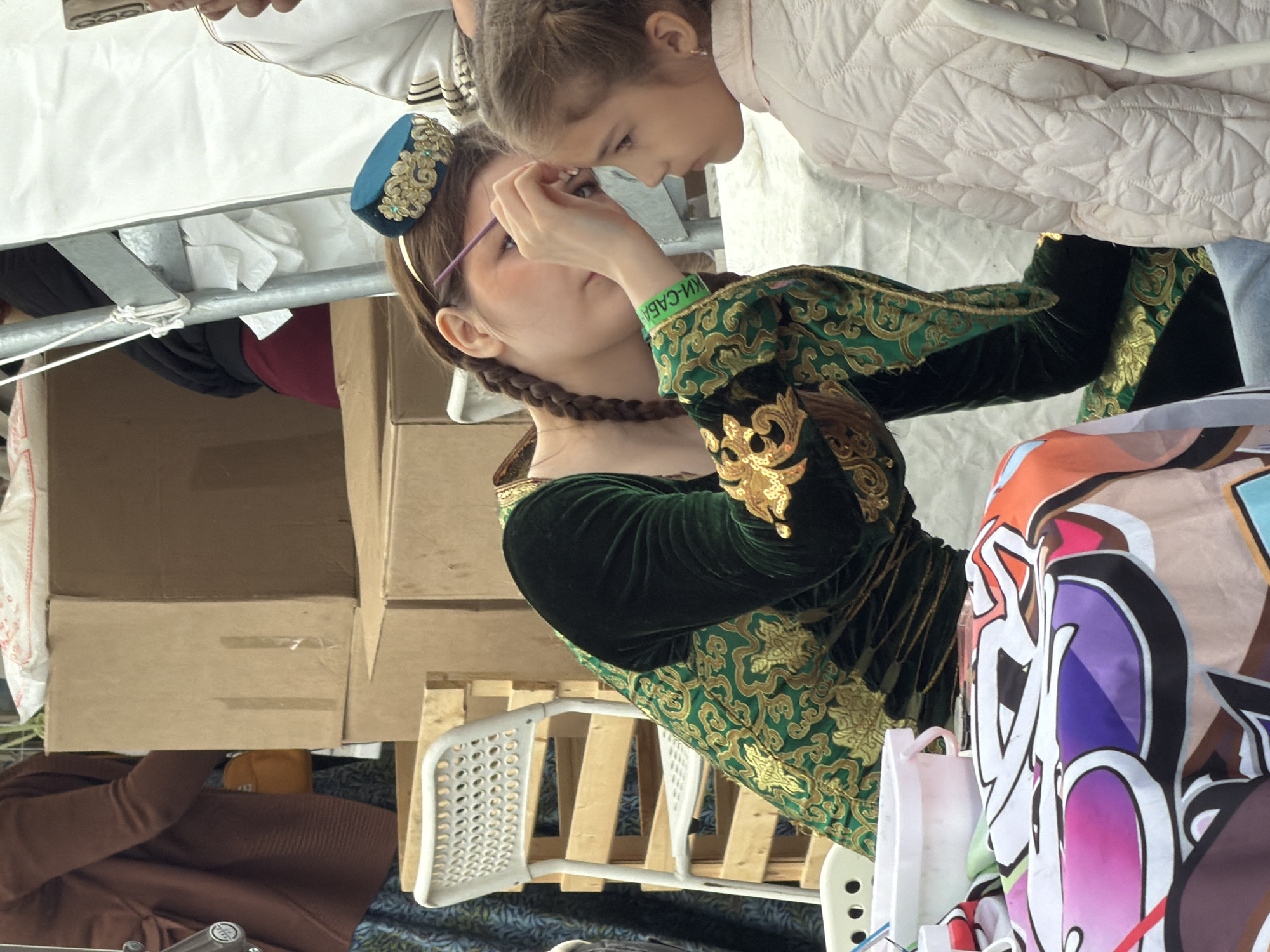 A young woman with braided hair, wearing a dark green velvet dress with gold trim, applies face paint to a young girl. The woman wears a small, ornate blue hat on her head. The young girl is wearing a light pink quilted jacket. They are seated at a table covered with a colorful cloth that has abstract designs. In the background, there are cardboard boxes and a white chair. The scene appears to be outdoors, possibly at a festival or market, with a white tent structure visible behind the people.