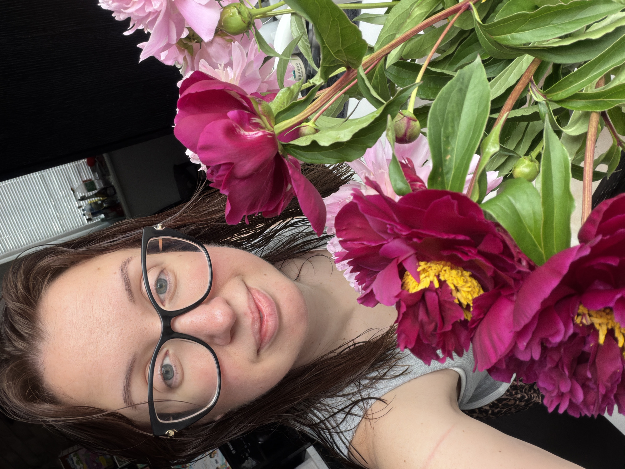 A medium shot captures a young woman with wet, shoulder-length brown hair, wearing black-rimmed glasses and a light grey tank top, holding a bouquet of pink and deep magenta peonies. She is looking directly at the camera with a slight smile, her head tilted to the left. The bouquet is positioned in the foreground, with the vibrant flowers partially obscuring her face. Behind her, the background is dimly lit and appears to be an indoor setting, possibly a room in Bila Tserkva, Ukraine, with a window covered by blinds and shelves with various items visible. The overall lighting suggests it is daytime, with the light source coming from the window.