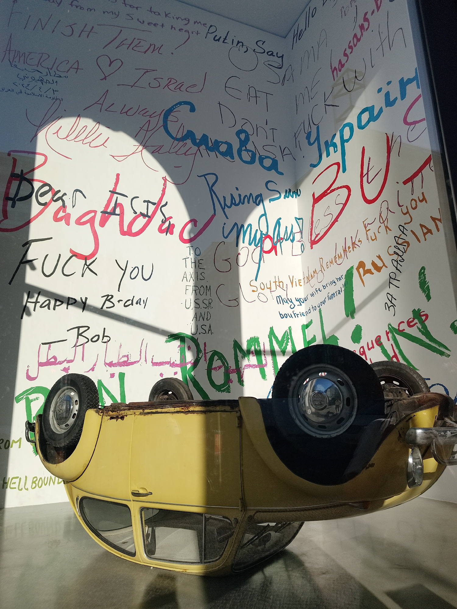A yellow Volkswagen Beetle is overturned in a storefront window, with graffiti scrawled on the white wall behind it. The graffiti includes words and phrases in various colors such as "Finish Them!", "America <3 Israel", "Dear Baghdad", "Fuck You", "Happy B-day Bob", "Ukraine", and some text in Arabic and Russian. The reflection in the car’s hubcap shows the surrounding environment.
