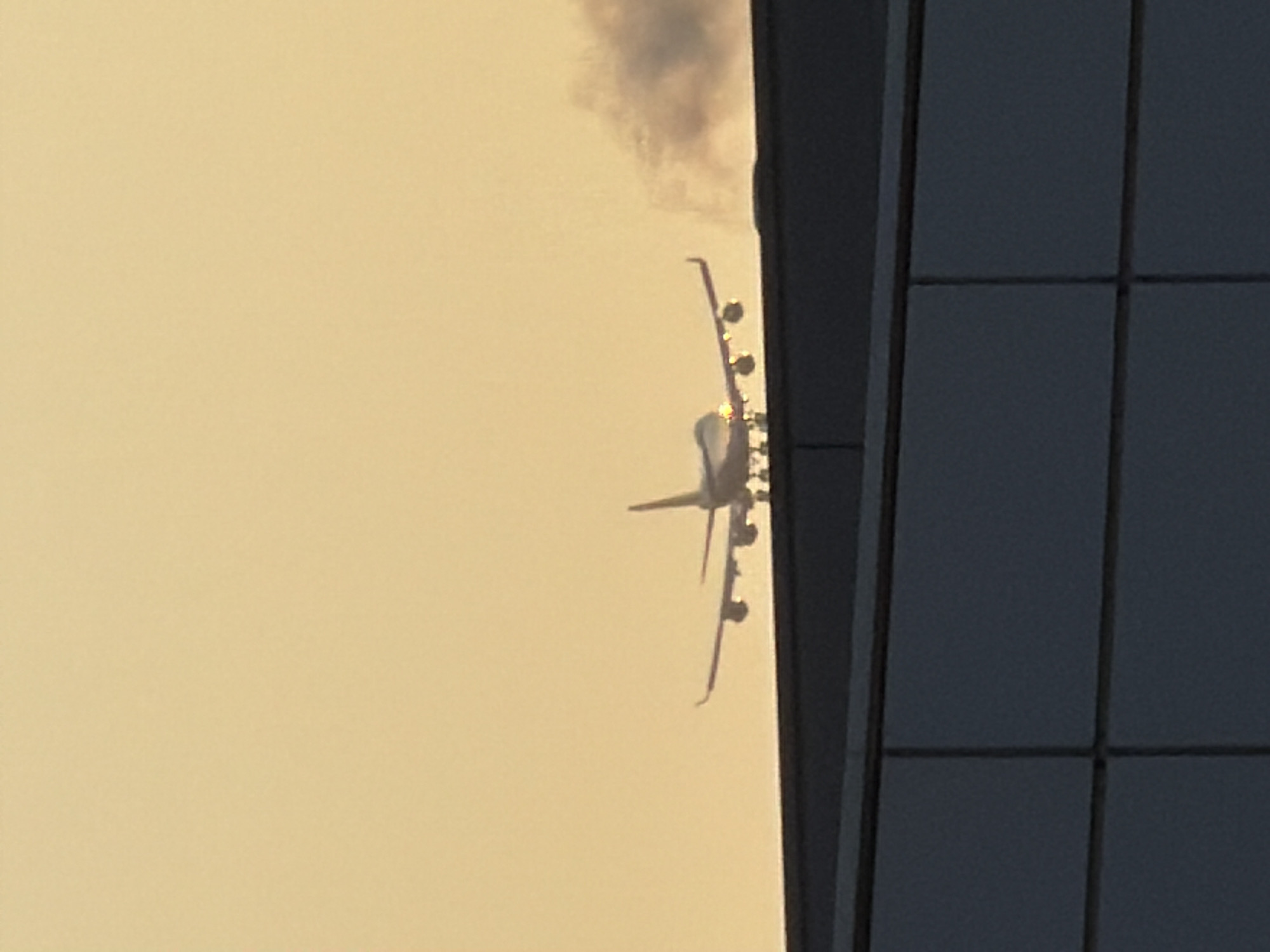 An airplane is captured mid-flight, silhouetted against a hazy, light-orange sky, likely during sunrise or sunset. The aircraft appears to be a large passenger jet, possibly a Boeing 747 due to its four engines and distinctive hump. It is flying low, with its landing gear extended, suggesting it is either landing or taking off. Smoke or exhaust plumes are visible trailing from the right side of the aircraft, and the airplane's lights are illuminated.
In the foreground, a dark, flat structure, possibly a building or a rooftop, extends horizontally across the bottom of the frame. This structure is divided into rectangular sections, suggesting it's part of a modern architectural design. The overall scene is viewed from a slightly elevated perspective, looking towards the aircraft. The location is the city of Leudelange, Luxembourg.