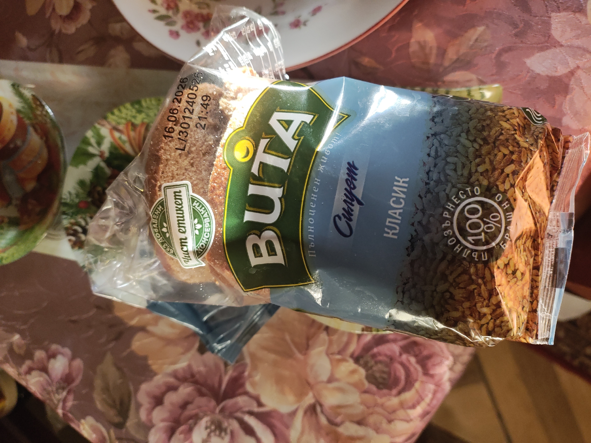 Here is a description of the image:
The image shows a package of "Vita" brand bread, "Siluet Classic" variety, on a table. The bread is in a clear plastic wrapper with a blue label that has the brand name "Vita" in large, green letters. Under the brand name, in smaller script, it says "Pylnotsenen zhiwot!" and "Siluet" in Cyrillic. Below that is "Klasik," also in Cyrillic. The package also indicates it contains 100% whole grain flour. The date "16.06.2026 L/301240526 21:49" is printed on the packaging. A label indicating “Clean Label - No added preservatives” is present in Bulgarian. The table has a floral pattern. There are dishes with food partially visible in the background.