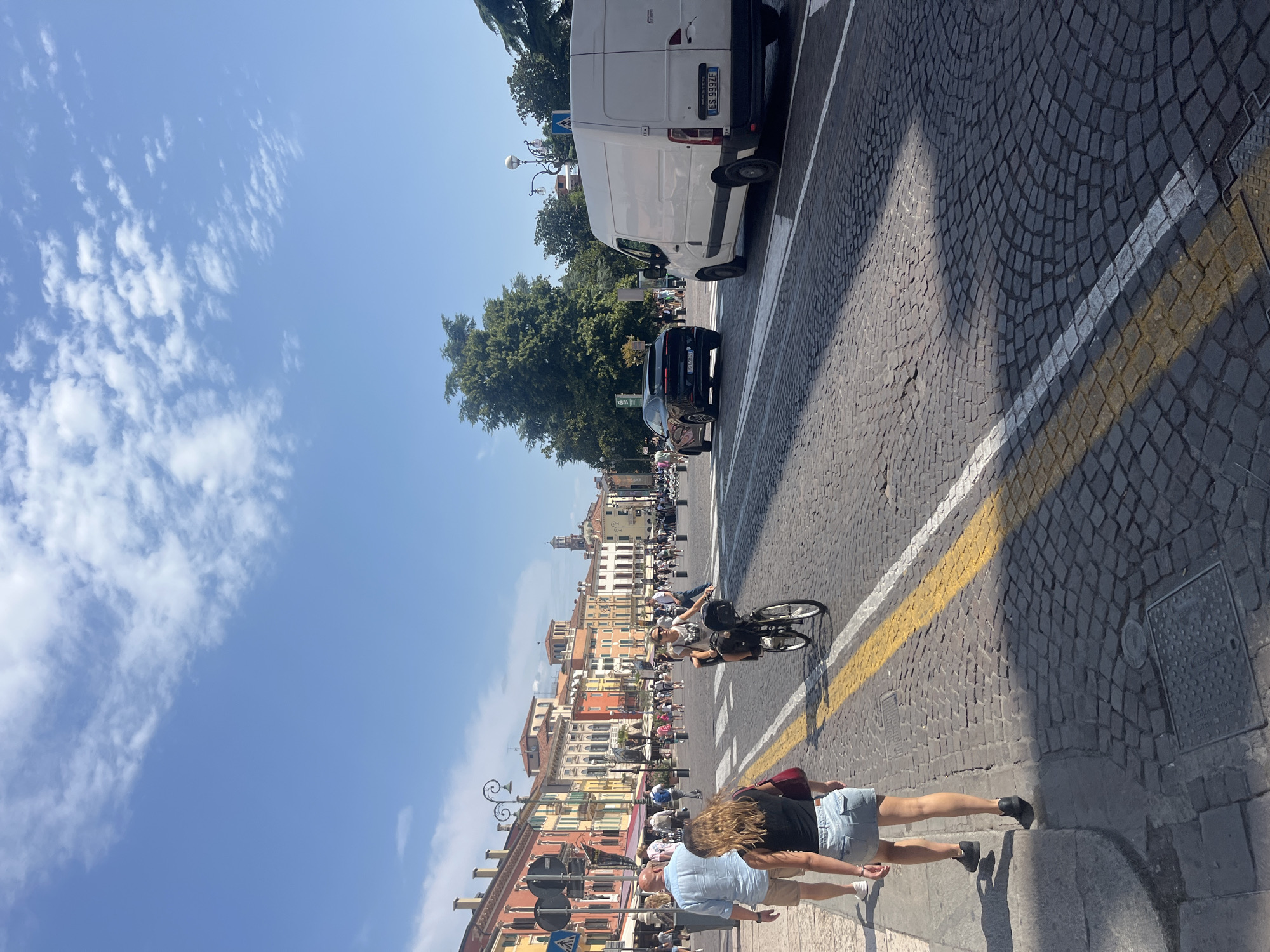 The image shows a view of Piazza Bra in Verona, Italy, on a sunny day. The piazza is paved with cobblestones. In the foreground, several people are walking on the sidewalk and crossing the street. A woman is riding a bicycle in the street, and a white van is parked on the right side of the street. In the background, there are several buildings with colorful facades, and trees. The sky is blue with some white clouds.