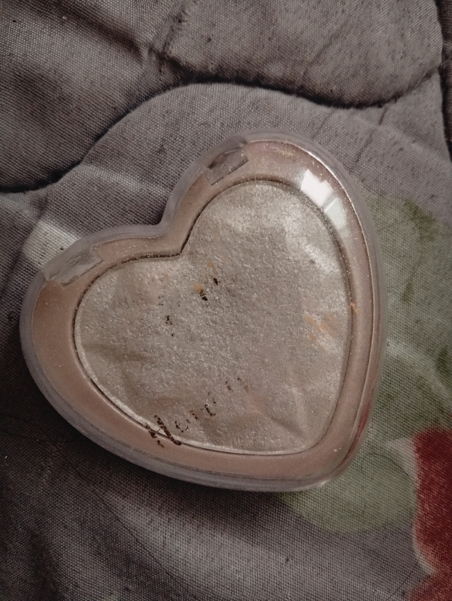A close-up image depicts a heart-shaped makeup compact, possibly containing highlighter or eyeshadow, resting on a textured, gray-toned fabric. The compact appears well-used, with visible wear on the surface of the makeup inside. Dark particulates are also embedded in the product. There are no identifiable people, specific actions, or immediate interactions visible in the image. The environment is restricted to the immediately visible surface texture, suggesting a fabric background. Location information, city of Poltava, Ukraine, cannot be determined from the image itself.