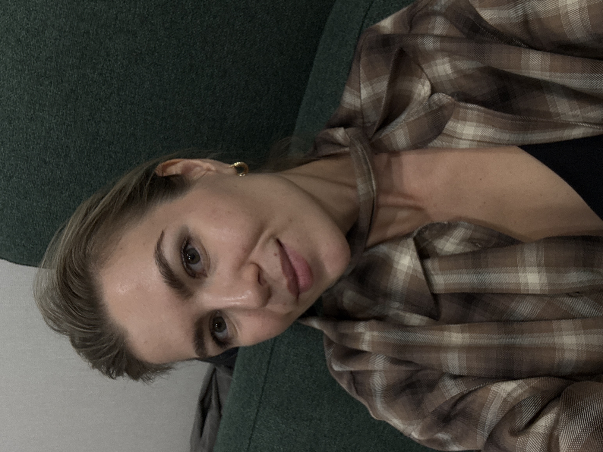 A person with blonde hair and light skin is seen from the chest up. The person is wearing a brown plaid shirt and a black top underneath. The background consists of a dark green textured surface, which could be a couch or chair. A small gold hoop earring is visible on the left ear. The location is Unknown, Unknown.