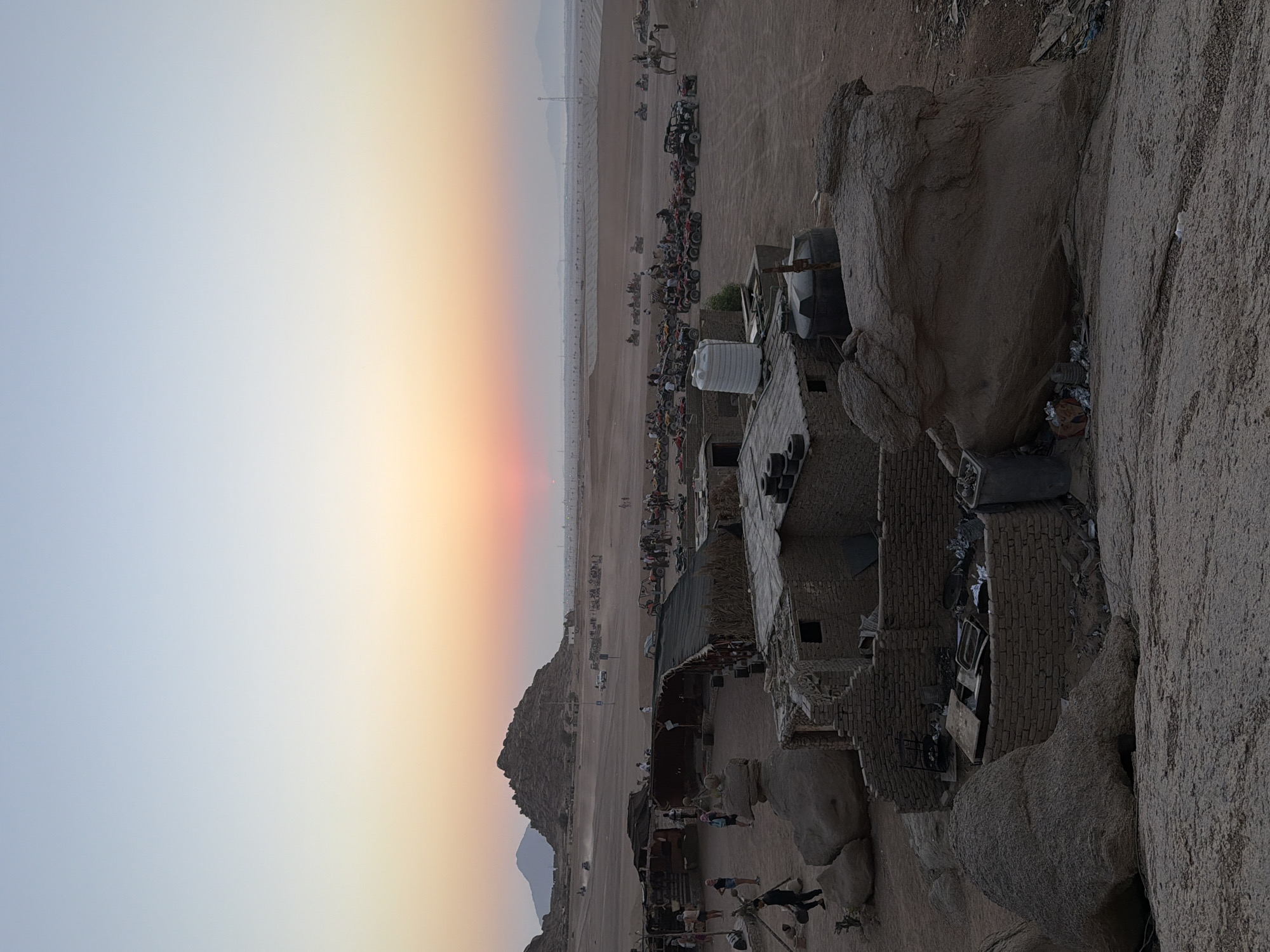 This is a photograph taken at sunset in Sharm El Sheikh, Egypt, likely in a desert area used for recreational activities. The sky is a gradient of pale blue and orange, with the sun a dim, reddish glow on the horizon. In the foreground, there are several rough, stone structures made of brick and corrugated metal, appearing somewhat dilapidated. Large boulders are scattered throughout the scene.
In the middle ground, there is a collection of ATVs and buggies parked in the sand, suggesting a rental or tour operation. A few people are visible, some standing near the vehicles, others walking around the structures. A camel stands near some of the ATVs, indicating a tourist attraction. Further in the background, more ATVs are seen in motion across the flat, sandy terrain. Distant mountains, hazy in the sunset light, form the backdrop. The environment is arid and dusty, characteristic of a desert landscape.
