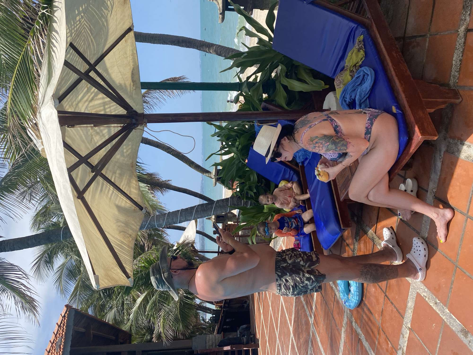 The image depicts a scene at a resort or beachside area in Phu Thuy Ward, Vietnam, on a clear, sunny day.
In the foreground, a Caucasian man stands facing left, positioned on a reddish-brown tiled patio. He is shirtless, wearing camouflage-patterned swim shorts, a dark green and brown plaid bucket hat, and white Crocs. He has a beard, sunglasses, and a tattoo on his right calf. His left hand holds a smartphone, which he is observing. To his right, a blue inflatable swimming ring lies partially visible on the tiles.
Adjacent to the man, to his right, three blue lounge chairs with wooden frames are arranged. The lounge chairs are positioned under the shade of a large, light-colored beach umbrella, the underside structure of which is visible.
A Caucasian woman is seated on the rightmost lounge chair. She wears a patterned bikini featuring floral and paisley designs in shades of purple, pink, and blue. She has extensive tattoos on her back and left arm/shoulder. She is wearing a straw fedora hat and sunglasses. Her left foot is bare with yellow-painted toenails, while her right foot, partially visible, wears a white sandal. She holds a halved tropical fruit, possibly a coconut, and appears to be consuming its contents. A blue towel and a patterned yellow/green towel are draped over her lounge chair.
Two young children are seated on the middle lounge chair. The child on the left, a boy, wears a blue long-sleeve swim shirt with "THE WAVE" text and blue swim shorts, along with blue swim goggles perched on his forehead. The child on the right, a girl, wears a light orange or pink short-sleeve shirt. Both children appear to be observing the standing man.
In the background, beyond the lounge chairs and lush green tropical foliage, a sandy beach leads to a body of calm blue-green ocean. Several small boats are visible in the distant water. Multiple palm trees with white wrapping on their trunks are scattered throughout the scene, and a building with a reddish-brown tiled roof is partially visible on the far left. The sky is clear and blue.
