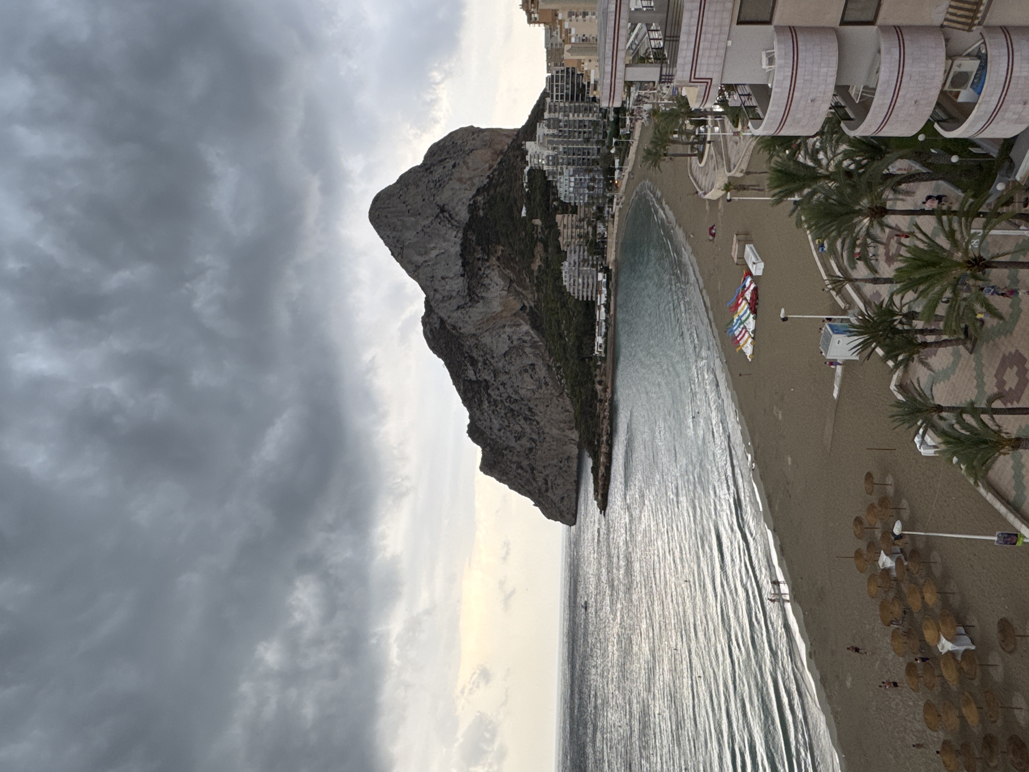This is an eye-level, wide shot of the coastline of Calp, Spain, on a cloudy day. The image captures a wide expanse of the ocean, a sandy beach, and a rocky outcrop that dominates the landscape. The water is a deep blue, with gentle waves lapping at the shore. The beach is populated with a few scattered umbrellas, lounge chairs, and a small group of people walking along the water's edge. In the background, a cluster of tall buildings lines the coast, creating a stark contrast with the natural beauty of the rocky outcrop. The sky is overcast with thick, dark clouds, creating a dramatic and moody atmosphere. The image seems to have been taken from a higher vantage point, possibly a balcony or a rooftop, looking down onto the beach and the sea.