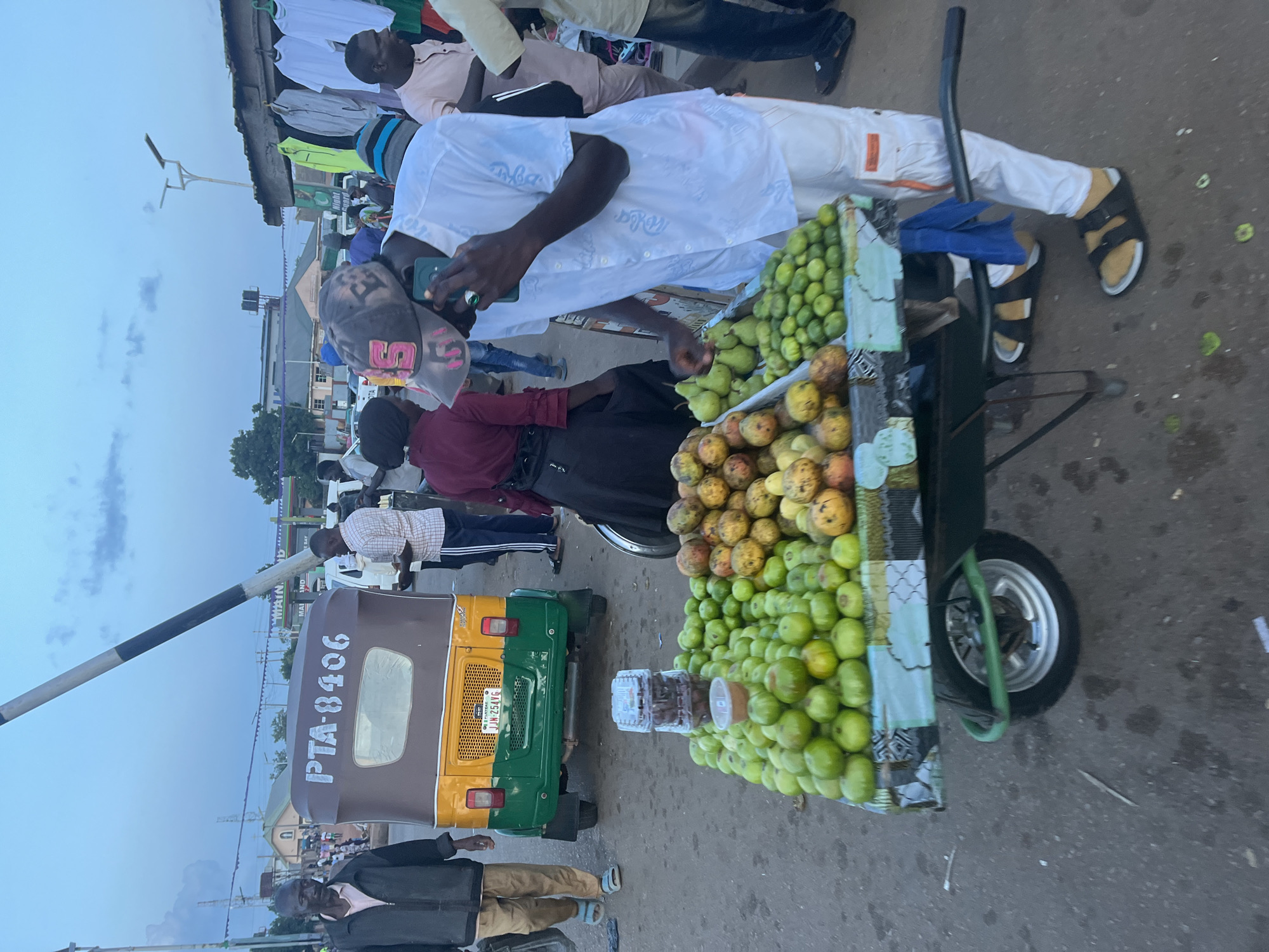 In Gura Top, Nigeria, a street scene unfolds at dusk. A man in a white shirt and white pants, wearing a baseball cap and black sandals, is on his phone while standing by a fruit cart. The cart is laden with green fruits, some yellow fruits, and several containers of dried goods. Behind the cart, a colorful auto-rickshaw with the license plate "PTA-8406" is parked. Several other people are visible in the background, some walking and others standing near market stalls that display clothing. The environment appears to be a busy market area with paved ground, overhead lighting, and a barrier arm extending across the street. The sky is overcast, suggesting the end of the day.
