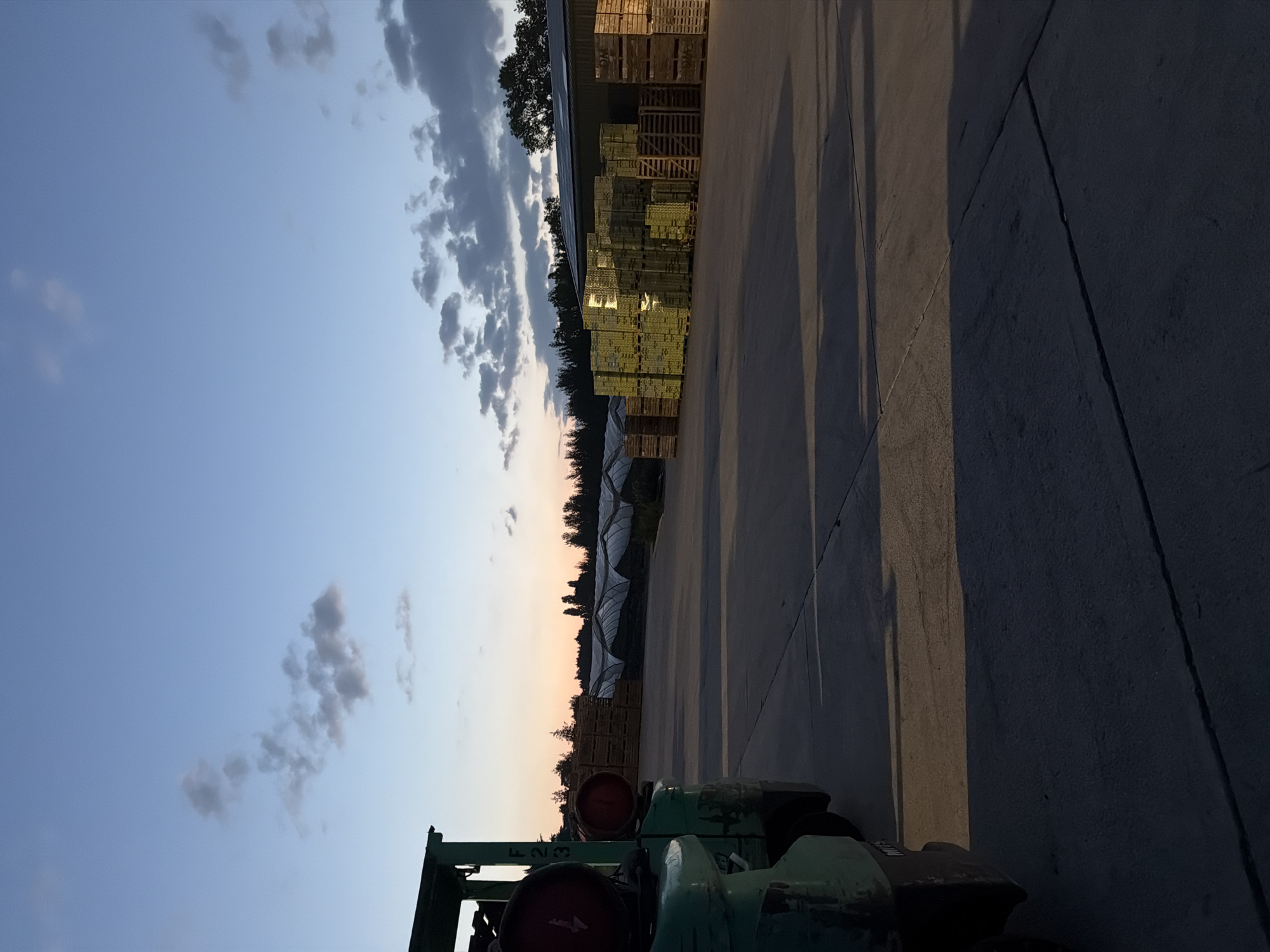This image captures an outdoor scene at dusk, likely in an industrial or agricultural area in Gravesham, United Kingdom. In the foreground to the left, a green forklift is partially visible, its lifting mechanism and a red gas cylinder prominent. The forklift is positioned on a concrete surface. The middle ground is dominated by a large, paved area, possibly a yard, with long shadows cast across it, indicating the low angle of the sun. To the right of the paved area, a substantial quantity of stacked wooden pallets is visible, some appearing to be filled with goods, possibly produce, and illuminated by the setting sun. Further back, rows of greenhouse-like structures with white coverings stretch into the distance, interspersed with trees. The sky above is a gradient of blue, transitioning to warmer tones near the horizon, with scattered clouds. The overall impression is one of an operational facility at the end of the day.