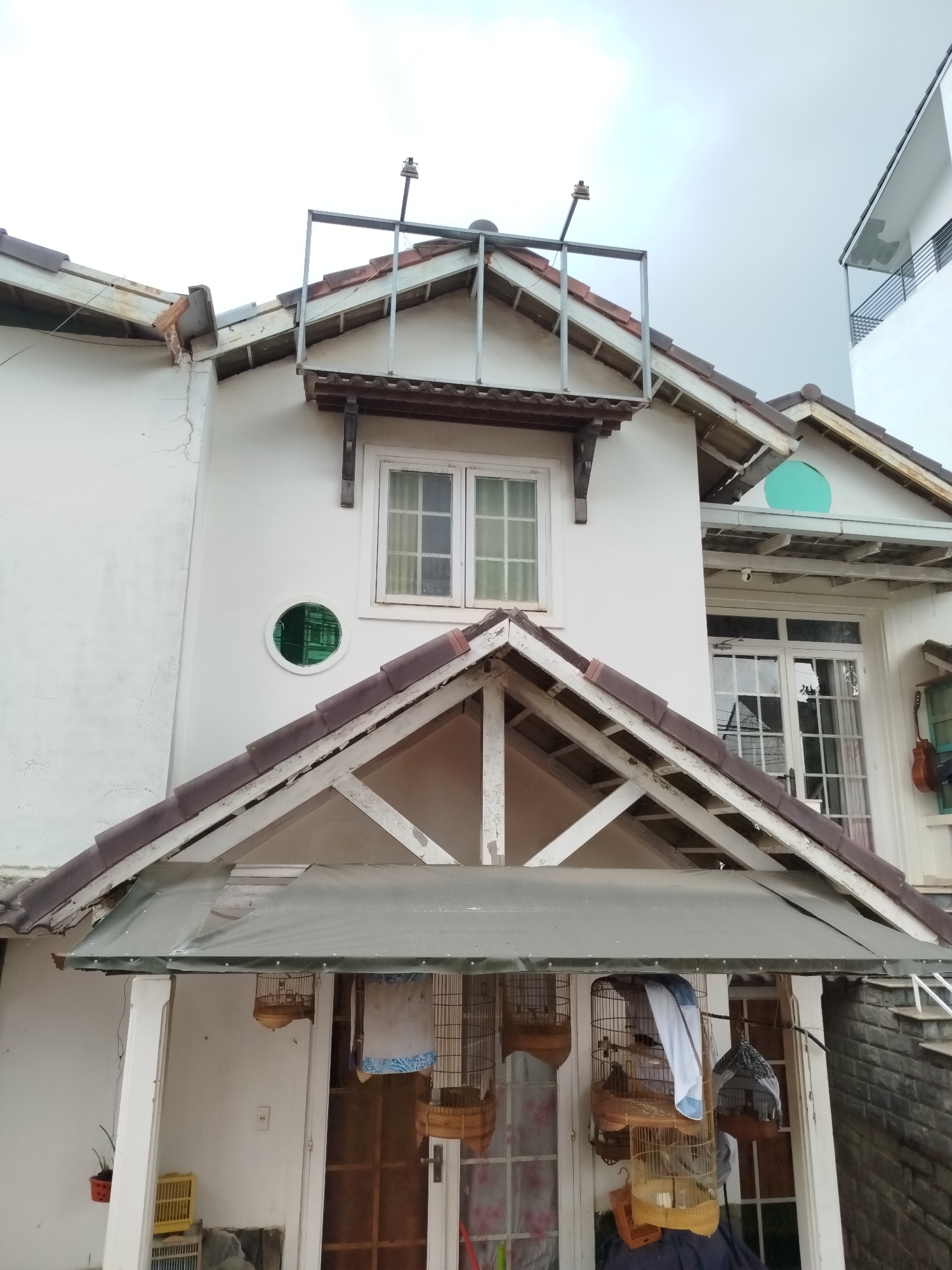 This image captures the exterior of a multi-level house in Da Lat, Vietnam, characterized by its traditional gabled architecture and worn, lived-in appearance. The primary subjects are the house itself, numerous bird cages, and an antenna-like structure on the roof.
The house features white-painted walls, which show some cracking and discoloration, and dark brown, possibly terracotta, roof tiles. Several prominent gables are visible, with exposed light-colored wooden rafters supporting the eaves. Windows vary in style: a large multi-paned window on the lower right, a smaller one directly above the main entrance, and a unique circular window with green-tinted glass on the upper left side of the facade.
Above the upper window, a metal frame structure, possibly for a sign or to support an antenna, is mounted on the gable. A slim pole with a small device, resembling an antenna or camera, extends upwards from this structure towards the overcast sky.
The activity suggested in the image is bird keeping, with multiple wooden and wire bird cages hanging or resting under a covered entrance area. Some cages are covered with white or patterned fabric. A white canopy extends over this entrance, supported by columns. To the right of the entrance, a guitar hangs on a wall. A small potted plant sits on a ledge to the left.
The setting appears residential, with another modern white building partially visible in the background to the far right. The sky is overcast, indicating daytime conditions but without direct sunlight. There is no visible text in the image. The overall impression is one of a functional, albeit slightly weathered, home.