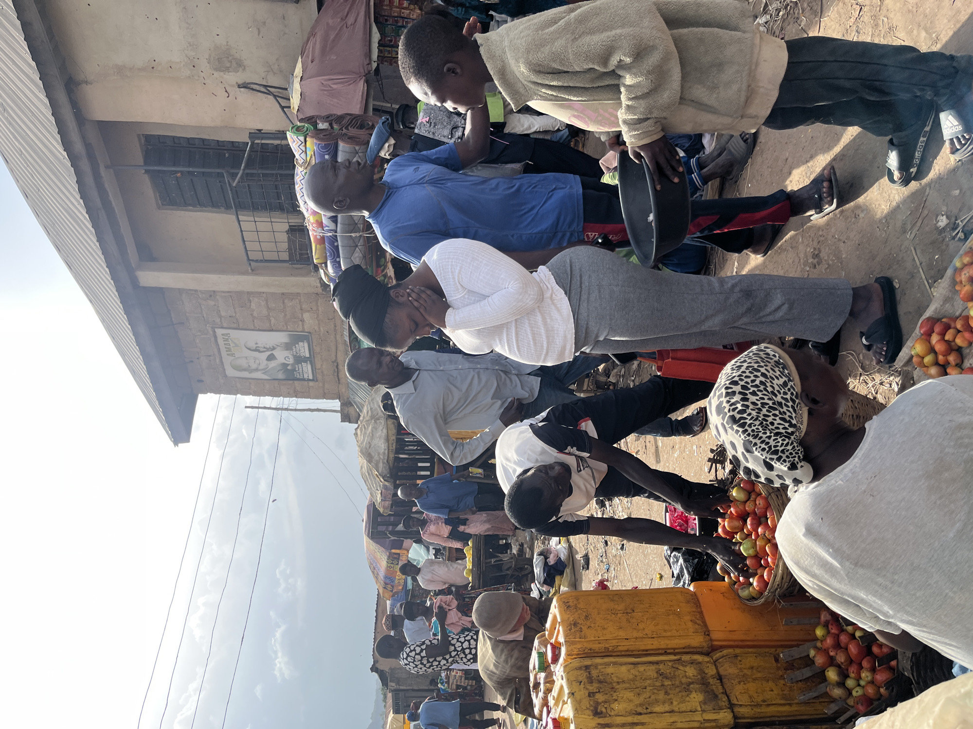 In Rafin Par, Nigeria, a bustling market scene unfolds. A pregnant woman in a white long-sleeved shirt and grey pants stands with her hand on her face, appearing distressed, next to a man in a blue t-shirt. A young boy in a beige jacket and dark pants holds a black bowl, looking towards the woman. Several other individuals are visible in the background, engaged in various market activities. Stalls with goods, including baskets of tomatoes and large yellow containers, are set up. A poster with images of two men is affixed to a building. The ground is dusty and uneven, typical of an outdoor market.