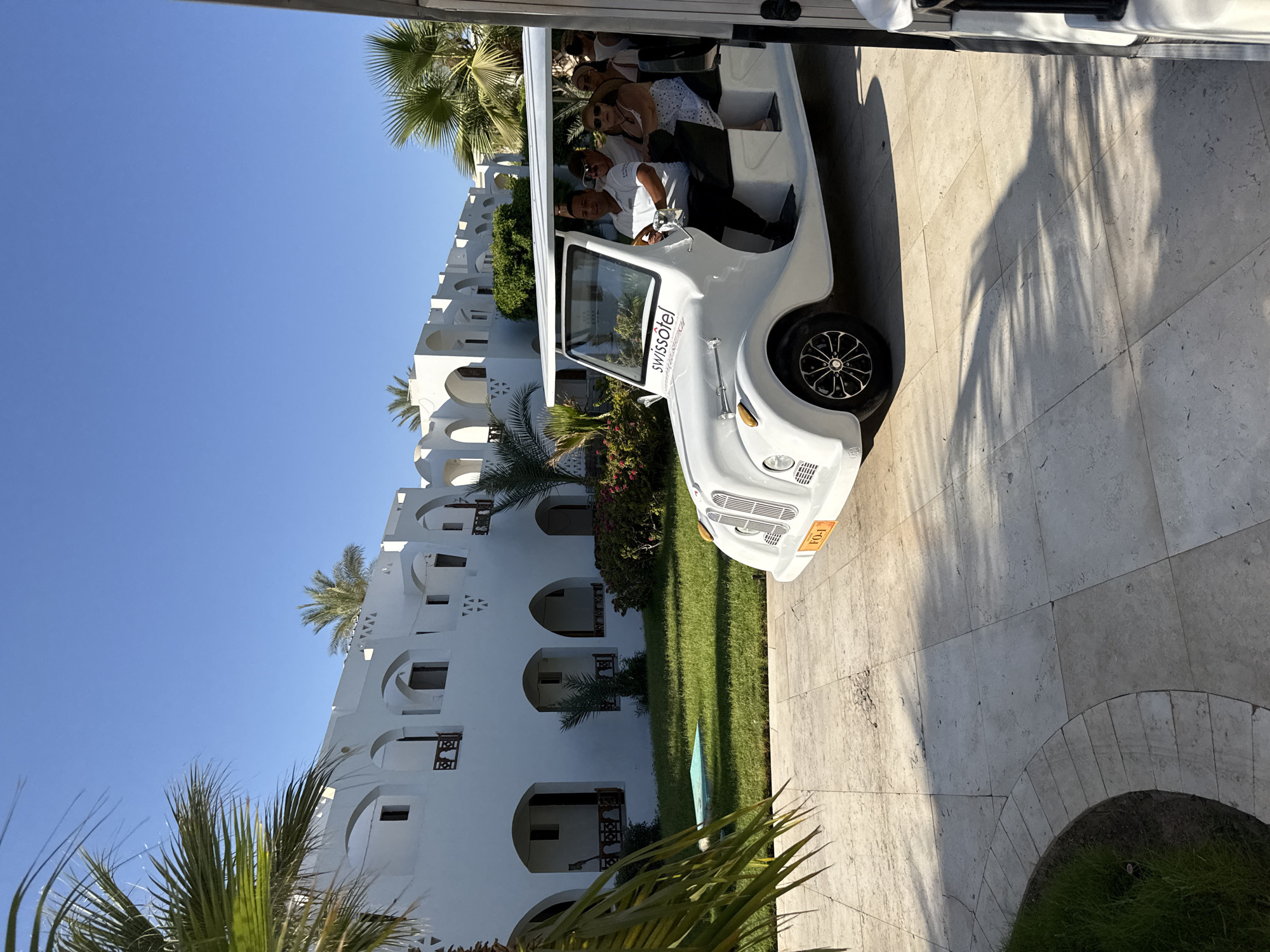 In Sharm El Sheikh, Egypt, a white electric golf cart labeled "swissôtel" is parked on a tiled pathway. The cart carries several passengers, including a male driver wearing a white polo shirt and sunglasses. Behind the cart, a white building with arched windows and balconies, typical of Egyptian architecture, stretches into the background. Palm trees are visible on the left side of the image, casting shadows on the pathway, suggesting a sunny day. The scene appears to be a resort or hotel property.