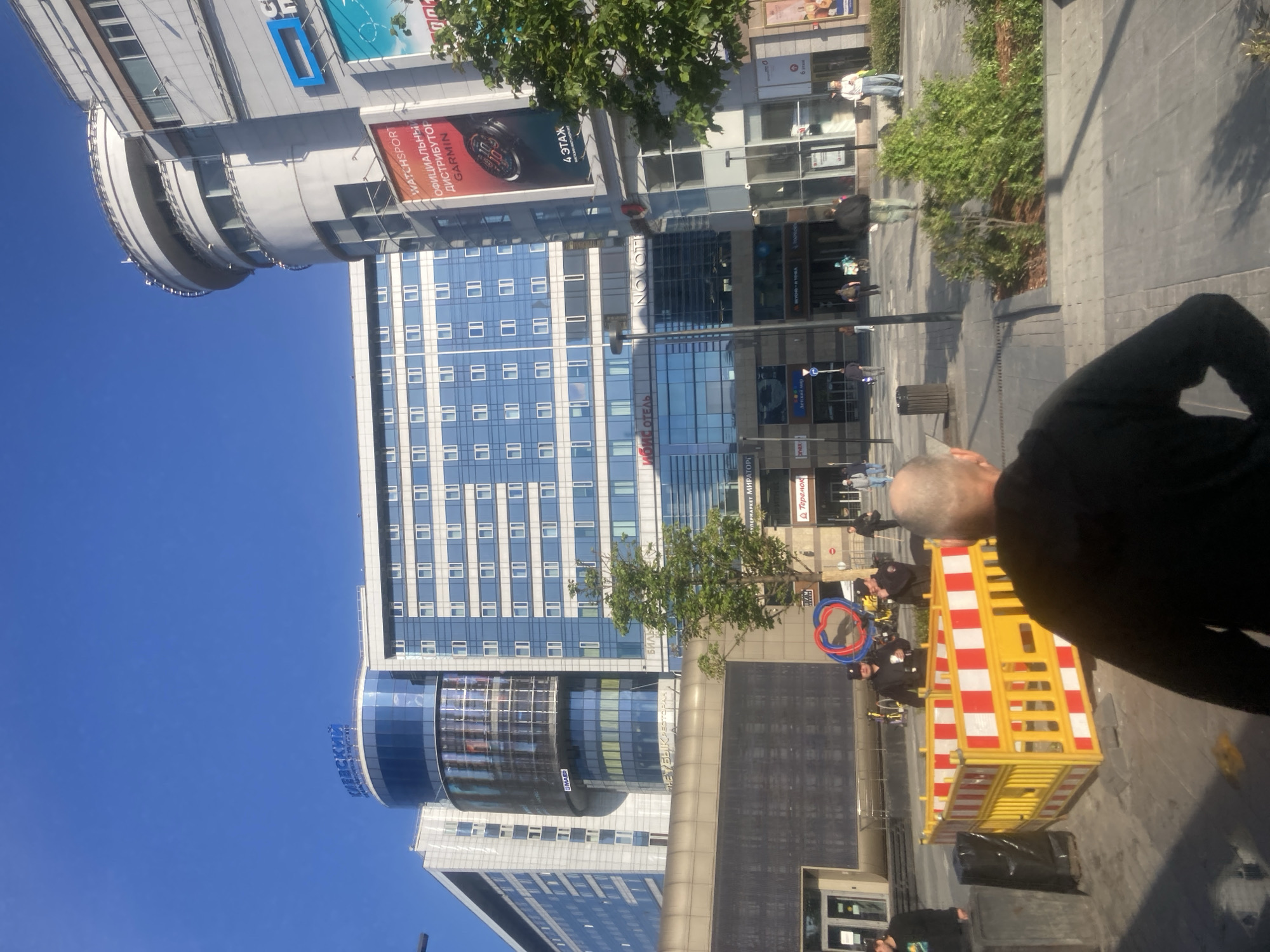 Here is a description of the image:
The image shows an outdoor scene in Moscow, Russia, featuring several buildings, including a tall hotel with a blue and white facade and a round section with the word "Киевский" on it, a Novotel, and a building with advertising, including one for "Watchsport" and Garmin. People are walking on the sidewalk. Some of them appear to be police near a yellow and red construction barricade.