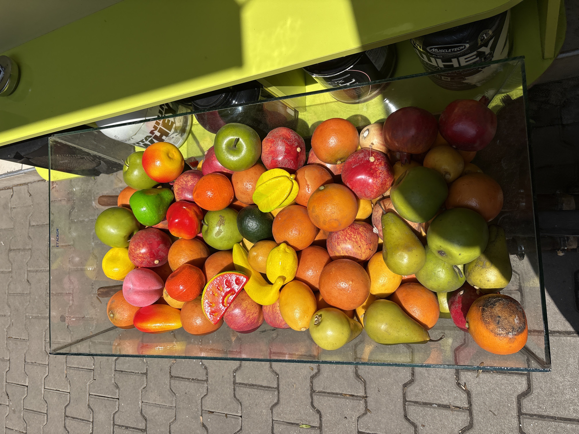 This image displays a vibrant collection of artificial fruits, tightly packed within a clear, rectangular glass container. The assortment includes a variety of green and red apples, oranges or tangerines, yellow and green pears, pomegranates, lemons, yellow bananas, a starfruit, and a slice of watermelon.
The glass container is situated outdoors on a paved surface composed of grey interlocking concrete blocks. Above the container, a bright yellow-green surface, possibly a countertop or part of a market stall, reflects strong direct sunlight, indicating a sunny day and casting distinct shadows.
Beneath the yellow-green surface, several dark, cylindrical containers are partially visible. One such container clearly displays the text "MUSCLETECH NITRO-TECH WHEY," suggesting it contains fitness supplements. Faintly inscribed on the top left edge of the glass container, the text "ET ACKOM" can be discerned.
The scene appears to be an outdoor display, possibly at a market, juice bar, or a similar commercial setting, with the artificial fruits serving as decoration or a visual representation of produce. No human activity is depicted.