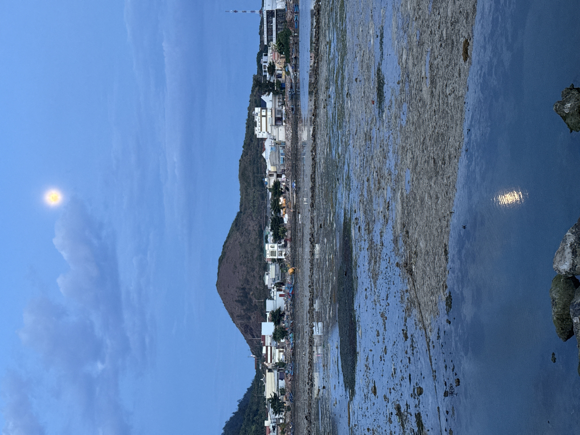 This image captures a coastal scene at dusk on Lý Sơn Island, Vietnam. In the foreground, shallow tidal pools reflect the twilight sky. The water is murky and appears to have a rocky, uneven bottom with some vegetation. A narrow strip of land, possibly a causeway or a rocky shore, separates the tidal pools from the main body of water.
In the middle ground, a line of small fishing boats is moored along the coast. Behind the boats, a village is nestled against the base of a rounded, vegetated hill. The buildings are a mix of low-rise structures, predominantly white and cream-colored, with some featuring pitched roofs. A few trees are scattered among the houses. A tall communication tower stands on the right side of the frame, near the edge of the village.
The background is dominated by the hill, which appears to be covered in sparse vegetation. The sky is a deep blue, transitioning towards darker tones, with wispy clouds. High in the sky, a bright, full moon is visible, casting a faint glow. The overall atmosphere is serene and suggests the end of a day.