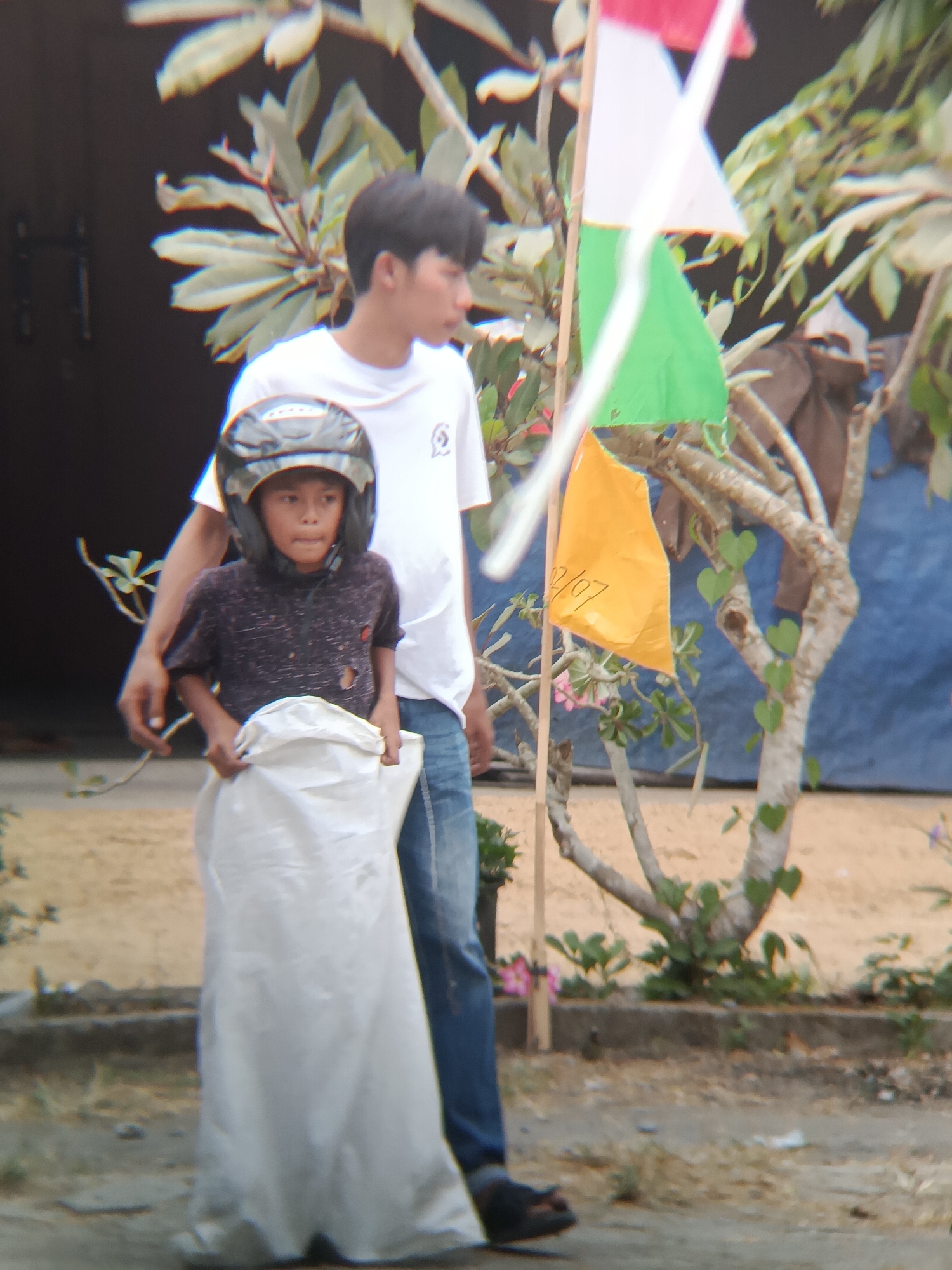 Two individuals are present in an outdoor environment, likely in Sarwadadi, Indonesia. A younger male, wearing a black helmet and a dark patterned shirt with a tear, stands in front of a larger individual. The younger male is holding a large white sack. The larger individual, wearing a white t-shirt and blue jeans, has an arm extended, seemingly supporting or guiding the younger male. Behind them, flags of red, white, and green, along with a yellow flag with writing that appears to be "02/07", are visible, attached to a wooden pole and partially obscured by foliage. The background consists of a dark structure, trees with green leaves and some pink flowers, and a blue tarp. The ground is a mixture of sand and dirt.