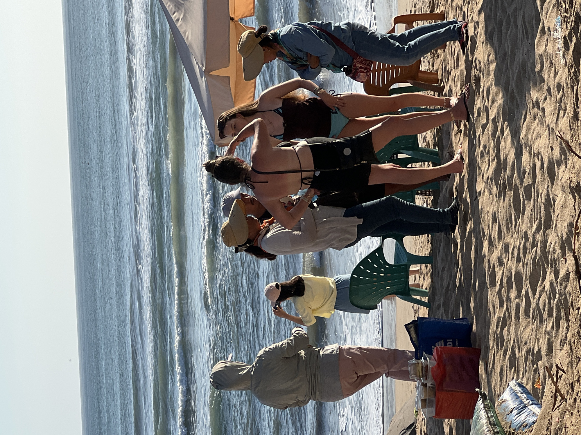 A group of people are gathered on a sandy beach in Kuta, Indonesia, near the ocean. The water is choppy with moderate waves, and the sky is clear.
Several individuals are standing and interacting. On the left, a person wearing a hooded jacket and pink pants stands facing the ocean, with a green plastic chair behind them. To their right, a person in a yellow shirt and blue skirt is taking a photo with a cell phone. Further to the right, a woman with a straw hat is conversing with another woman in a black bikini and shorts. A woman in a brown top and blue bikini bottoms stands between them, and on the far right, a person in a blue denim jacket and jeans is standing.
In the foreground, there are several objects on the sand, including red and blue bags, plastic containers with food and drinks, and some other items that are not clearly identifiable. There are also two chairs, one green plastic chair and one brown plastic chair, positioned near the group of people and a light-colored umbrella. The sand is textured with footprints and small debris. The overall scene suggests a casual gathering or outing on the beach.
