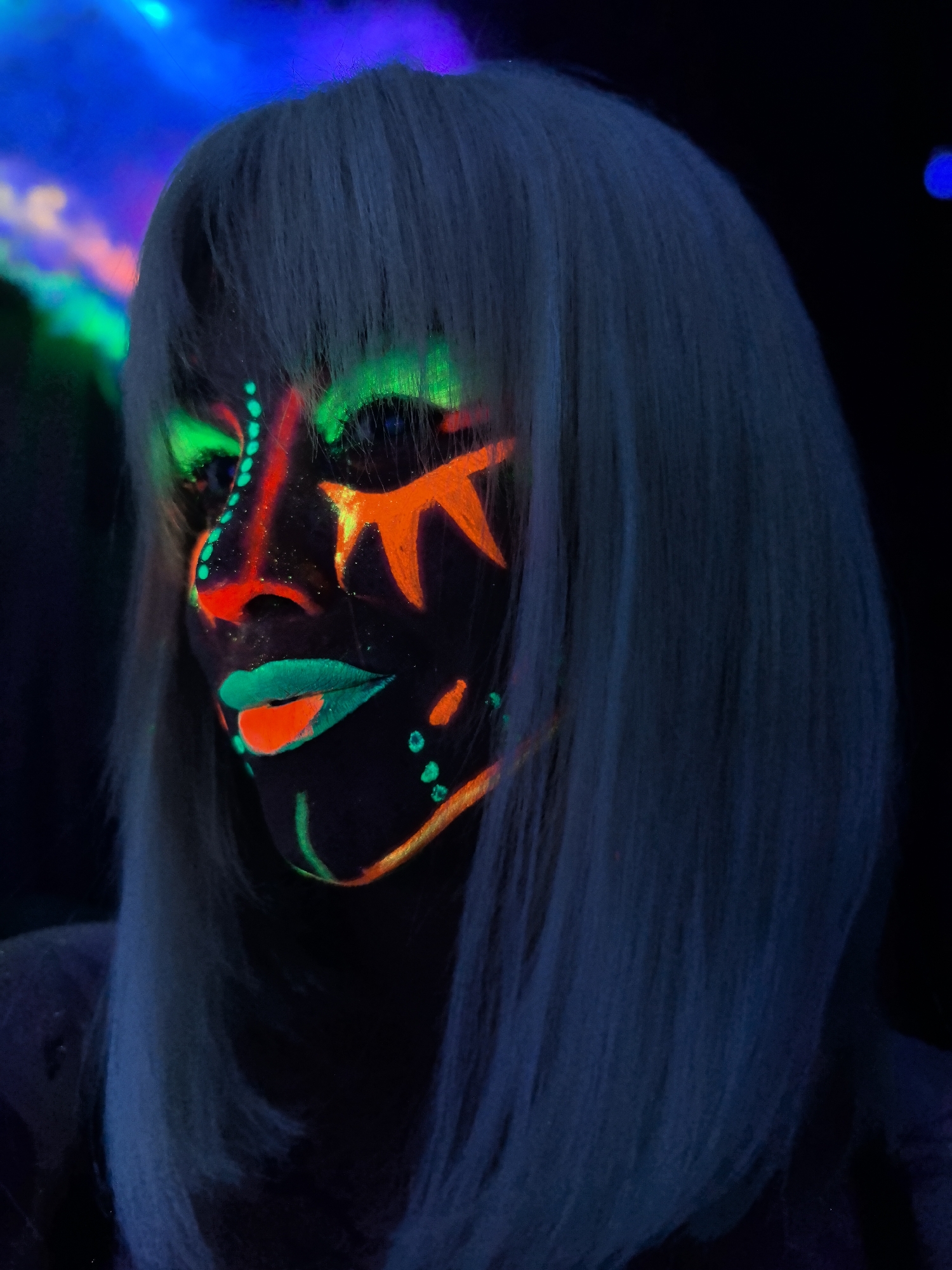 A person with light-colored, shoulder-length hair, possibly a wig, is captured in a close-up portrait, viewed from the upper chest up and angled slightly to their left. Their face is intricately painted with glow-in-the-dark or UV-reactive makeup, illuminated brightly by blacklight.
The makeup features vibrant neon green around the eyes and forehead, transitioning to red and orange lines and dots on the bridge of the nose and cheekbones. The lips are painted a striking teal or mint green, outlined with bright red. Geometric patterns, including starbursts and scattered dots in orange and yellow, adorn the cheeks and jawline. The skin beneath the makeup is very dark, contrasting sharply with the luminous colors.
The setting is an indoor, dark environment, strongly suggesting the use of UV or blacklight. The background is blurred but shows intense washes of blue, green, and hints of pink/purple light, indicating additional colorful lighting effects. The overall atmosphere is vibrant and artistic, typical of a glow party, rave, or a themed photoshoot. The person is actively posing for the photograph. No specific textual elements are visible. The time of day is clearly night or an artificially darkened interior, and weather is not a factor.