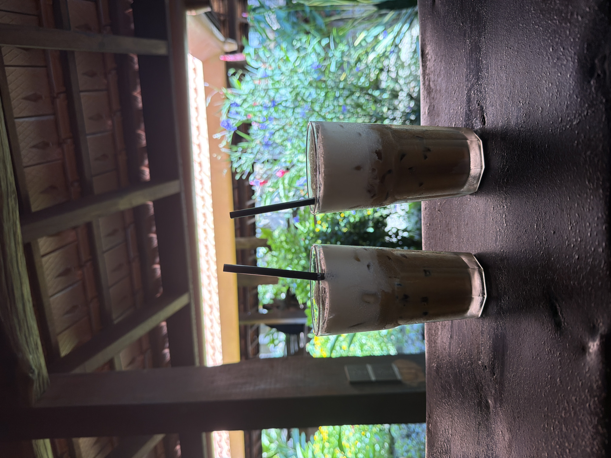 The image shows two tall glasses of iced coffee with milk foam on top, each with a black straw, placed on a dark wooden surface. The glasses are in focus, while the background is blurred, showing lush green foliage with hints of colorful flowers. Above the foliage, there is a wooden roof structure. The scene appears to be outdoors, possibly at a cafe or restaurant with a garden setting in Tay Nha Trang Ward, Vietnam. There are no people clearly visible in the foreground or midground, though indistinct figures can be seen in the background.