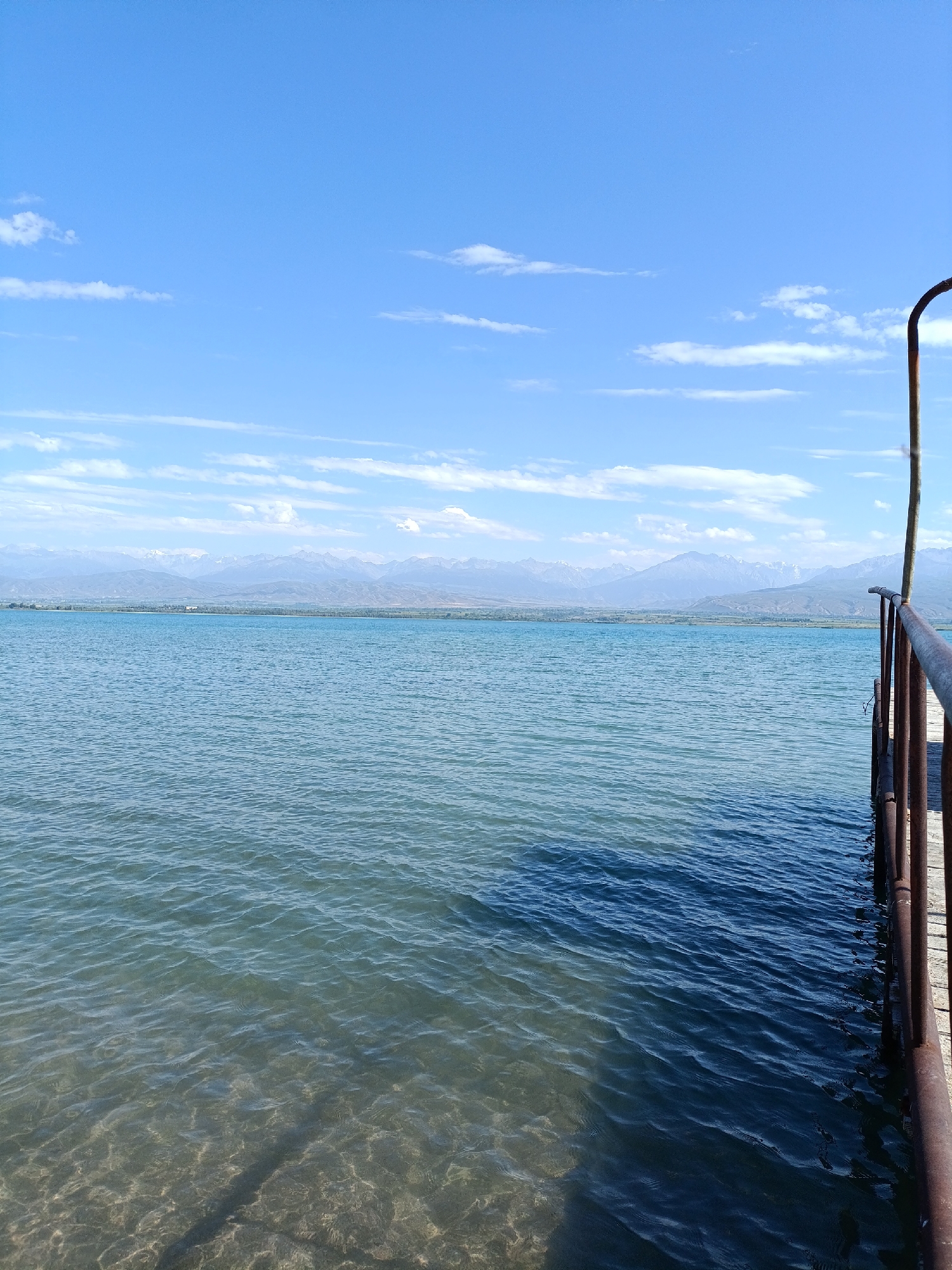 A wide-angle, eye-level shot captures a serene lake under a vast, bright blue sky dotted with scattered white clouds. In the foreground, the clear, shallow water reveals a sandy bottom, gradually deepening and darkening further from the shore. A wooden pier extends into the lake from the right side of the frame, its weathered planks and rusty metal railing indicating age. The pier casts a prominent shadow on the water's surface. In the distance, a range of majestic, snow-capped mountains forms a dramatic backdrop, separated from the lake by a strip of green land. The scene is peaceful and untouched, suggesting a natural, outdoor setting likely located in or near Kirpichny, Kyrgyzstan, given the mountainous terrain and clear water. No people or animals are visible in the image.