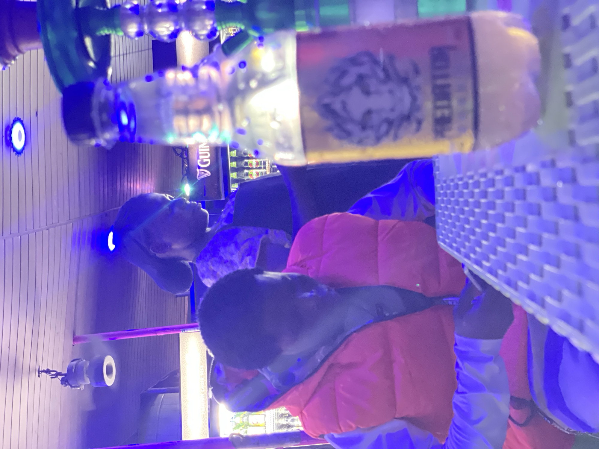 In Ado, Nigeria, the image depicts a dimly lit indoor establishment, possibly a bar or lounge, bathed in the vibrant glow of blue and purple neon lights. In the foreground, a clear plastic bottle with a yellow label featuring a predator's face and the word "PREDATOR" is placed on a white woven table, its surface illuminated by the ambient lighting.
Behind the bottle, a person wearing a bright orange puffer vest over a light-colored long-sleeved shirt is seated at the table, their face obscured by shadow as they look down, possibly interacting with a device or engaging in conversation. To the left, a refrigerator unit displays several bottles of Guinness.
In the background, another person wearing a dark cap and a patterned shirt is visible, their head turned to the side. The ceiling features a wooden panel design, with a hanging light fixture and a circular blue light fixture mounted overhead. The overall ambiance suggests a social gathering or entertainment venue.