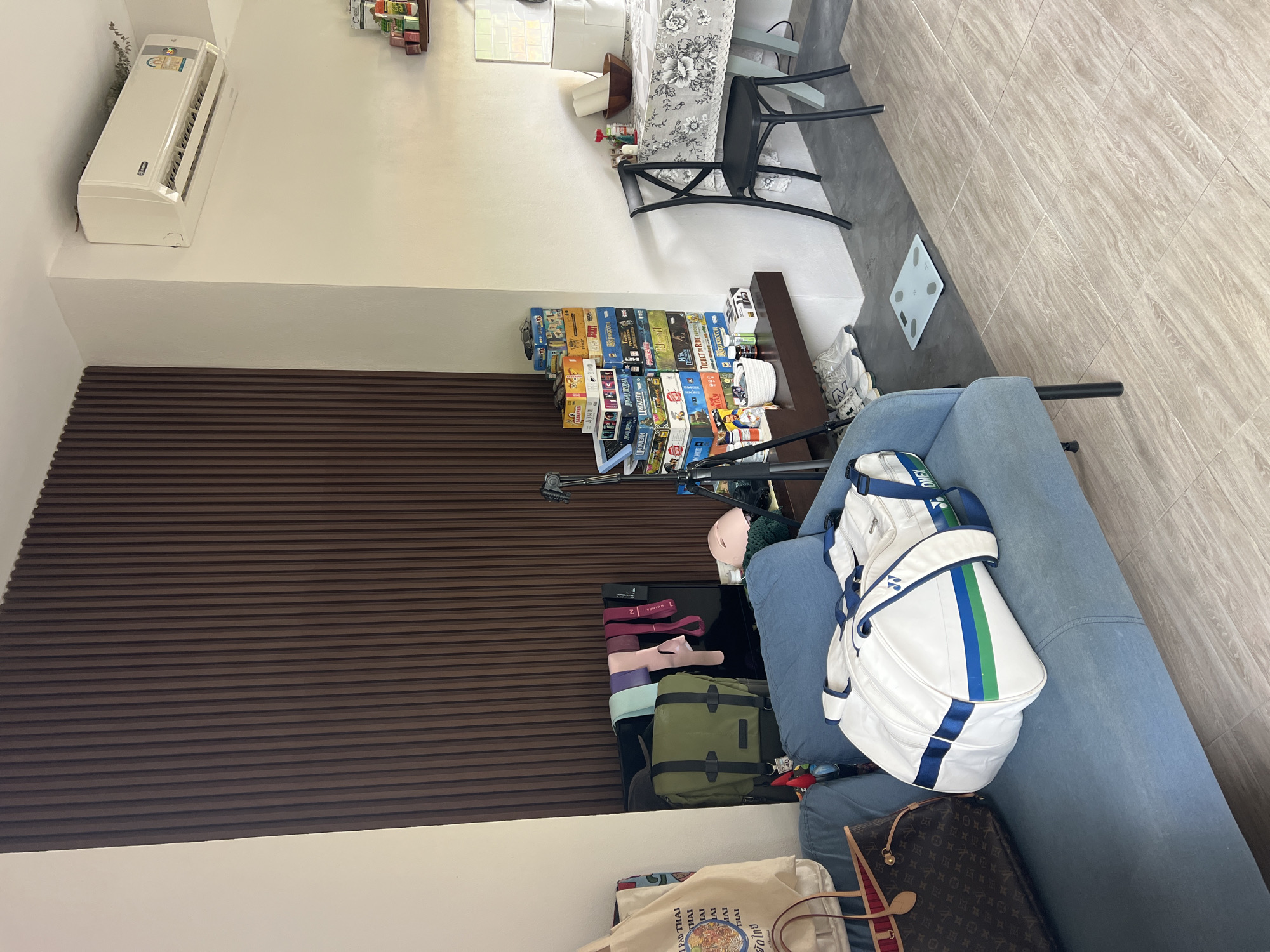The image shows a cluttered interior space in what appears to be a residential building. The perspective is slightly angled, giving a wide view of the room.
In the upper left corner, an air conditioning unit is mounted on the wall. Below this, a dark brown wooden wall panel extends downwards. A stack of board game boxes sits atop a dark brown table, with various colors and titles visible.
A light blue sofa occupies the right side of the image, on which lies a white golf bag with blue and green stripes, along with a brown Louis Vuitton handbag. Several other items, including a tote bag, are partially visible beneath the sofa.
Behind the sofa, a digital scale rests on the floor. In the distance, a dining table with a floral tablecloth is visible, accompanied by chairs. The room's flooring has a beige tile appearance with wood grain patterning.